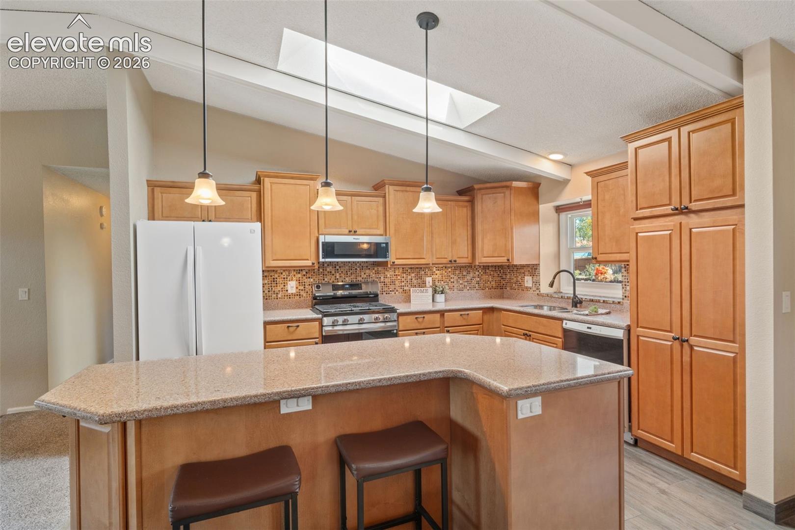 Kitchen featuring natural wood-finish cabinetry, stone-texture countertops, a mosaic tile backsplash, a vaulted ceiling with a skylight, and three pendant lights