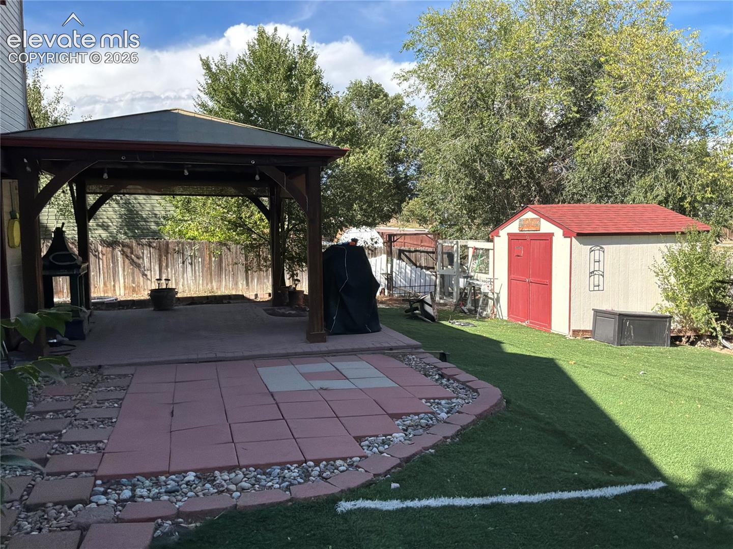 Covered patio structure with a concrete pad, a separate paver patio area, and an artificial turf lawn