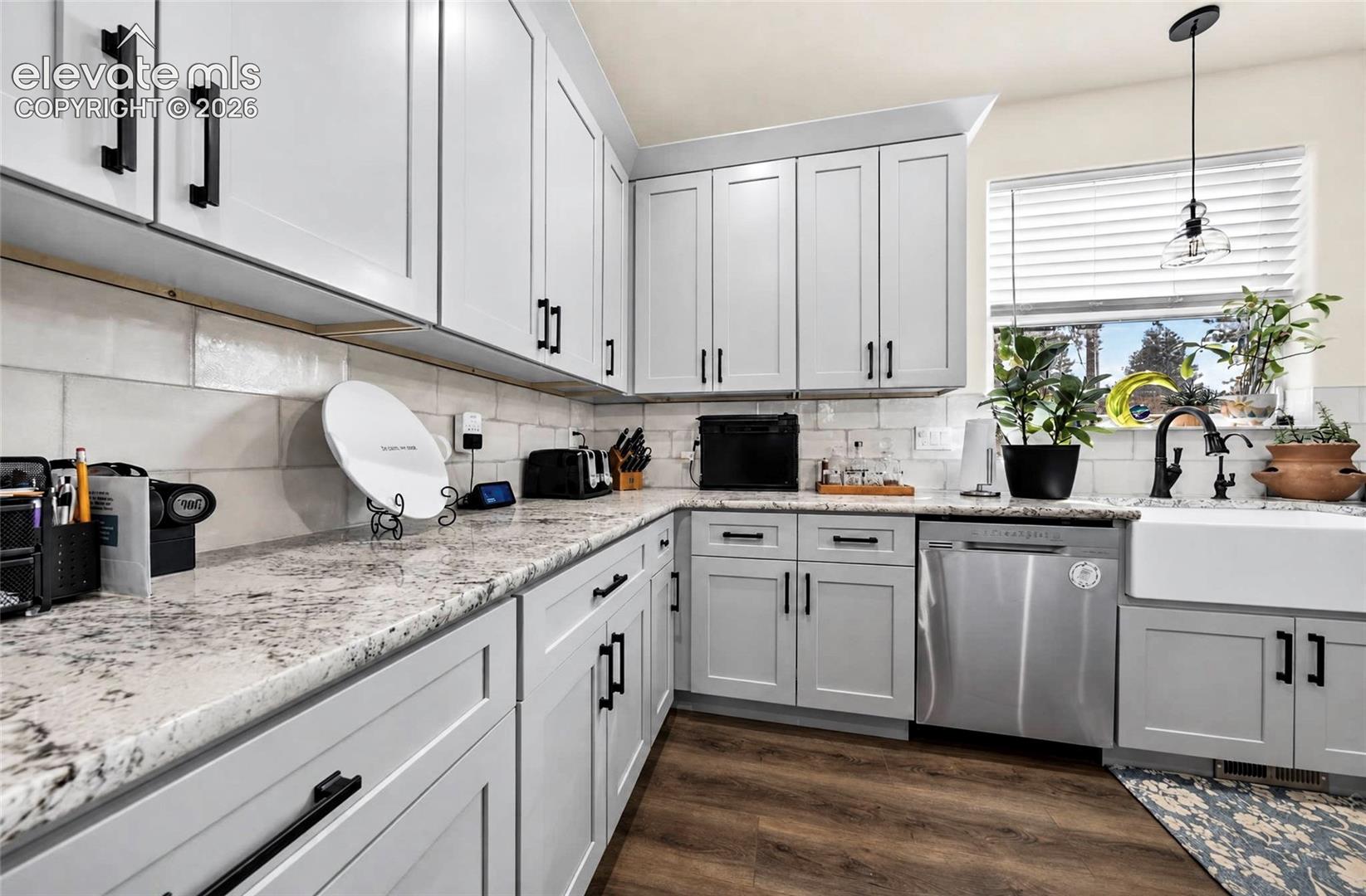 Kitchen featuring light gray shaker-style cabinetry, speckled stone countertops, a white farmhouse apron-front sink with a matte black faucet, subway-style wall tile backsplash, and wood-finish flooring