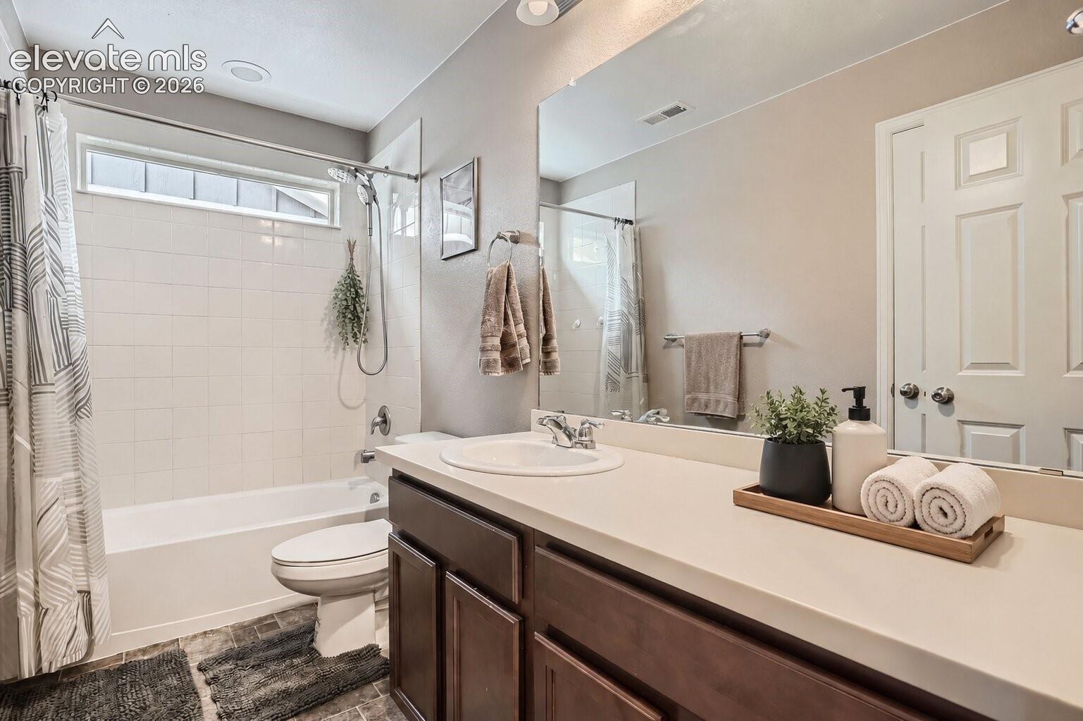 Bathroom featuring a single-sink vanity with dark wood-finish cabinetry, a solid-surface countertop, a full-width wall mirror, and a tile-surround tub-shower combination with an integrated transom window