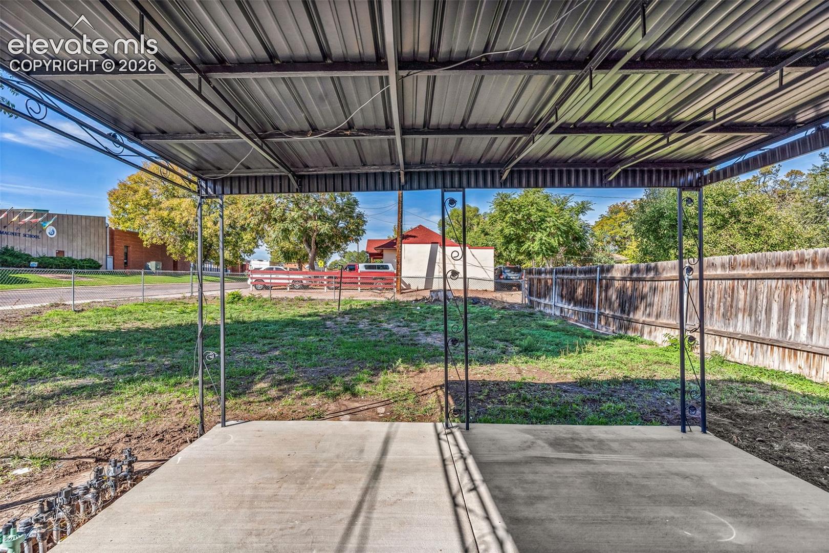 Covered outdoor concrete patio featuring a metal roof structure with decorative iron support posts and wood-plank perimeter fencing