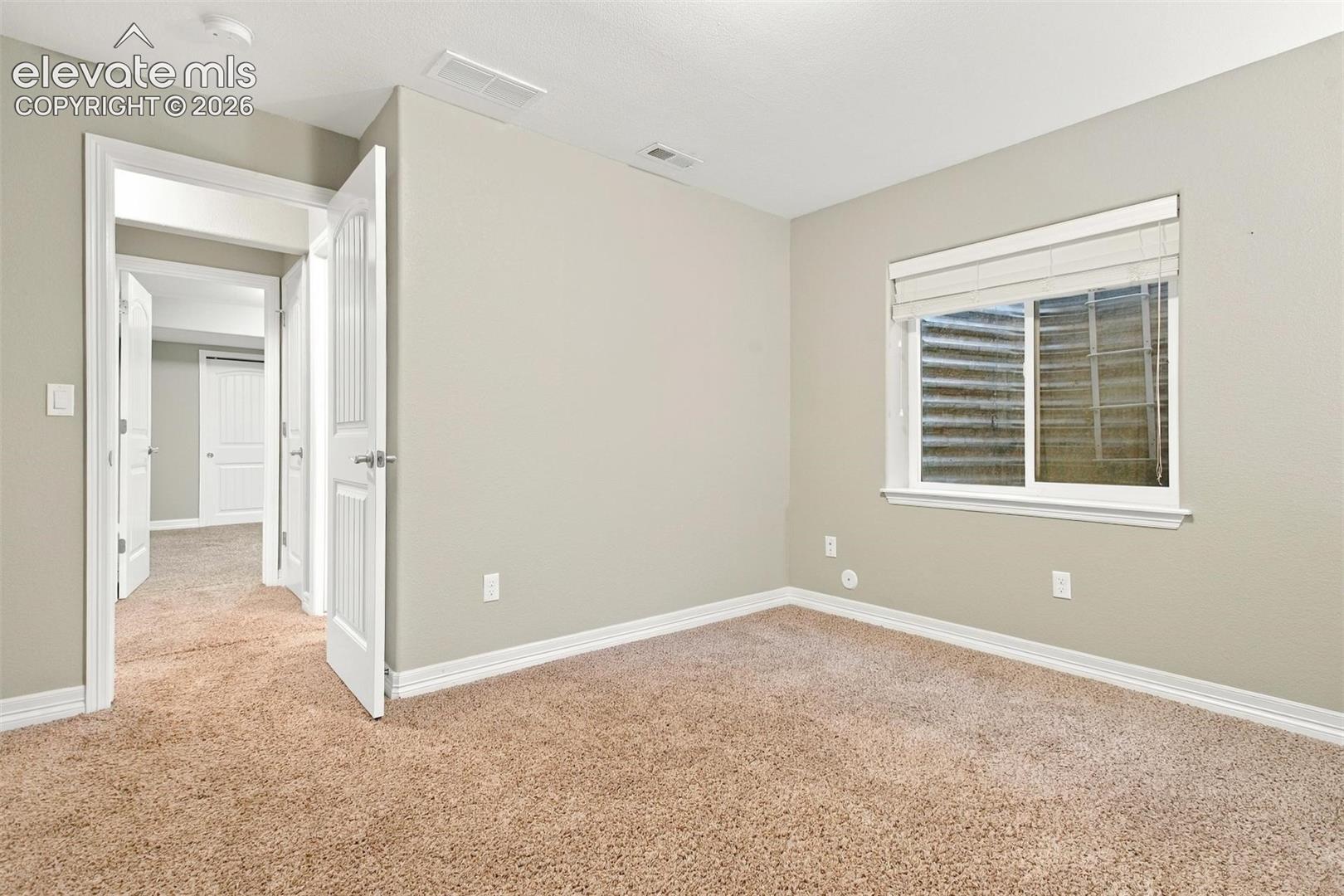 Neutral wall-toned interior featuring wall-to-wall carpeting, white paneled interior doors, and a sliding window with a corrugated metal window well