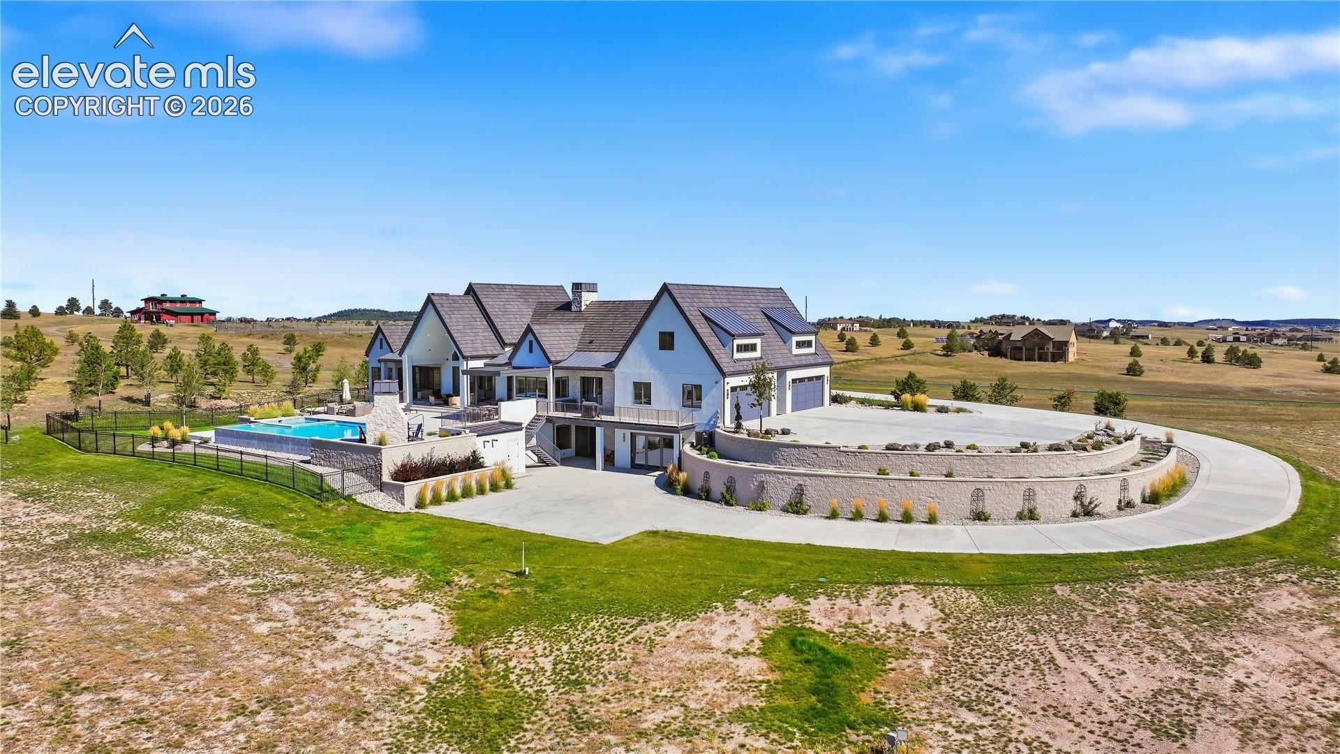 Multi-level residence featuring a dark shingle roof, integrated solar panels, concrete pool, tiered masonry retaining walls, and a circular concrete driveway