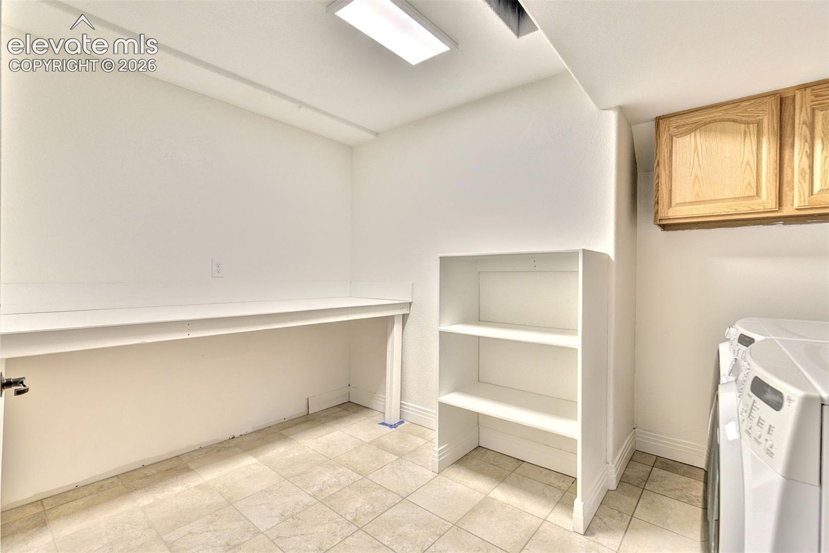 Utility room featuring tile flooring, built-in white shelving, an integrated workstation counter, and overhead wooden cabinetry