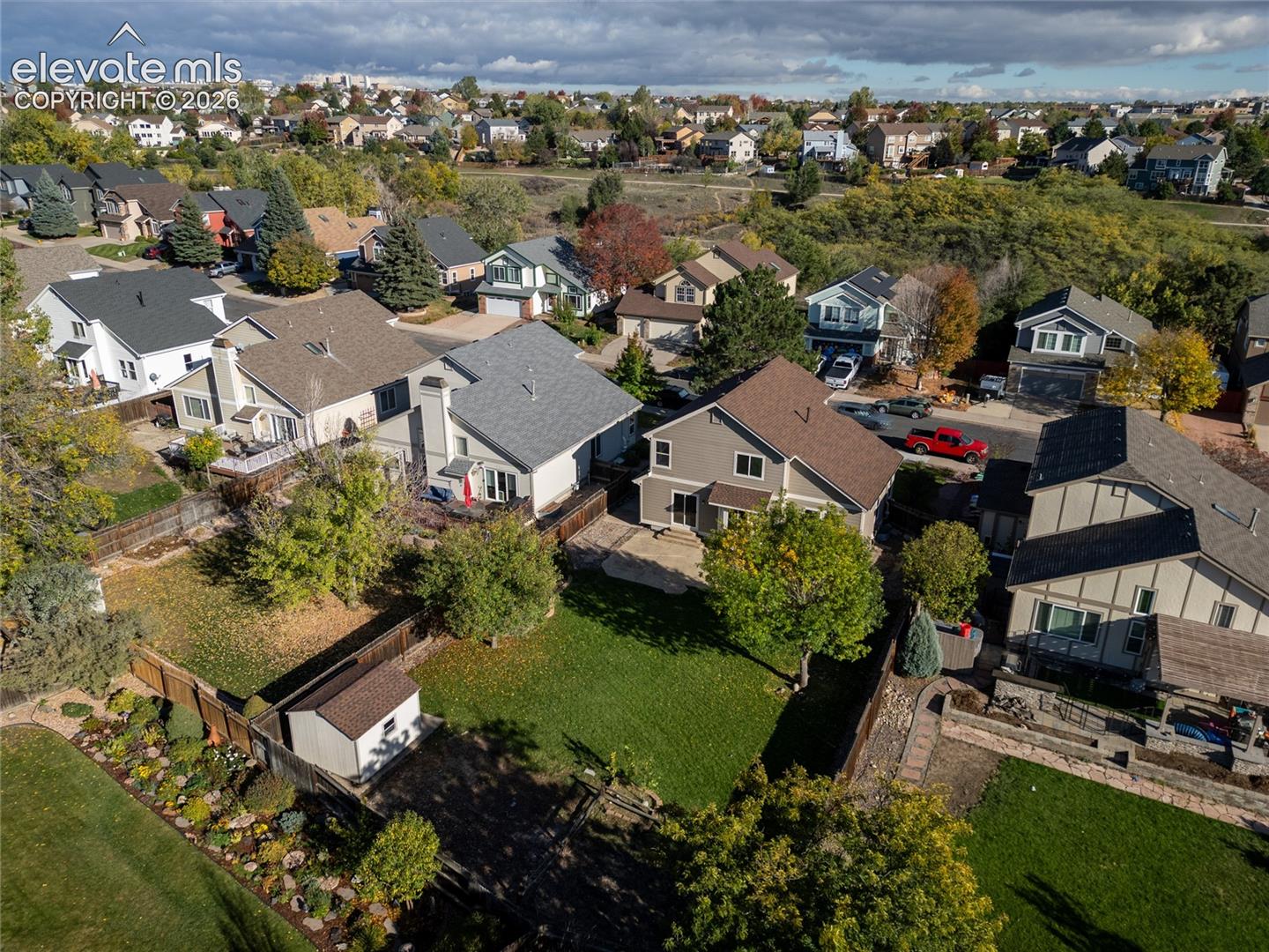 Aerial perspective of a multi-story residence featuring a paved rear patio, fenced backyard, detached storage shed, and mature trees