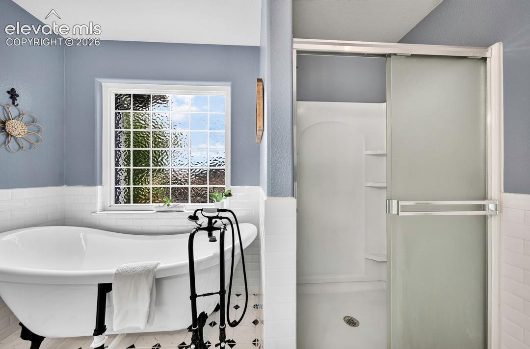 Freestanding clawfoot tub with matte black floor-mounted faucet, separate shower enclosure with sliding glass door, white subway tile wainscoting, and patterned tile flooring