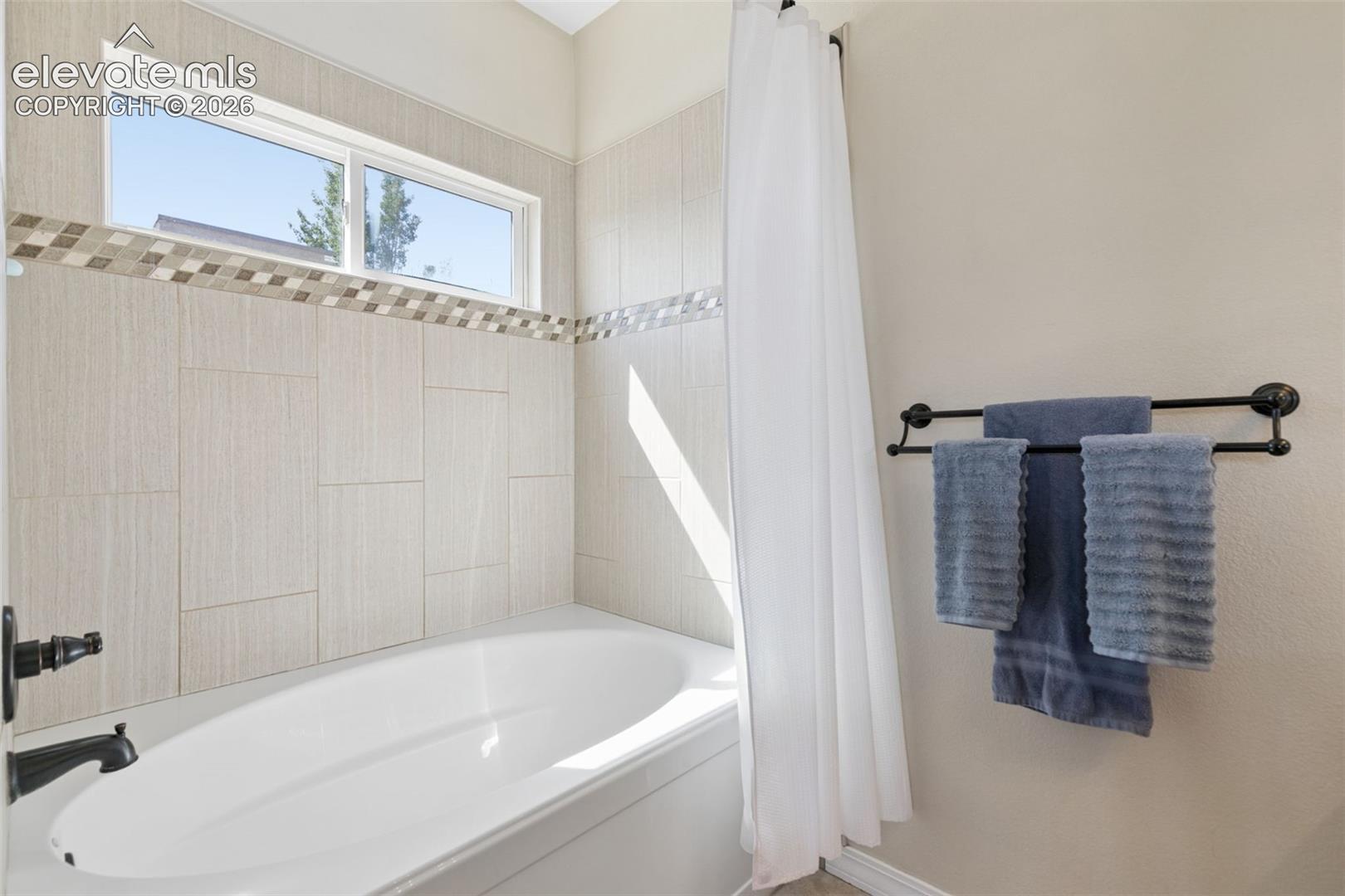 Built-in bathtub with a white finish, tiled surround with mosaic accent, and a single window