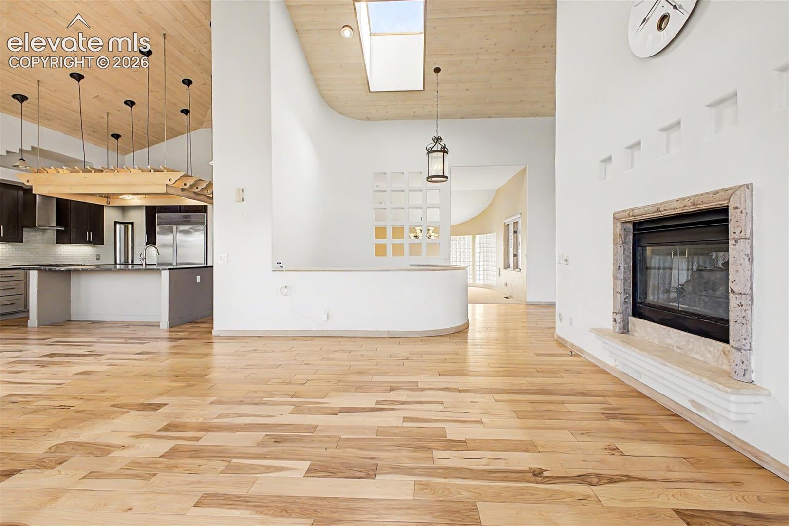 Hardwood flooring throughout, a stone-framed fireplace, a skylight, a kitchen island with dark cabinetry, and pendant lighting