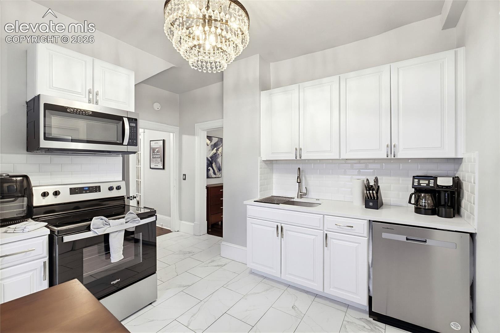 White shaker-style cabinetry, stainless steel appliances, white subway tile backsplash, white countertop, and crystal chandelier
