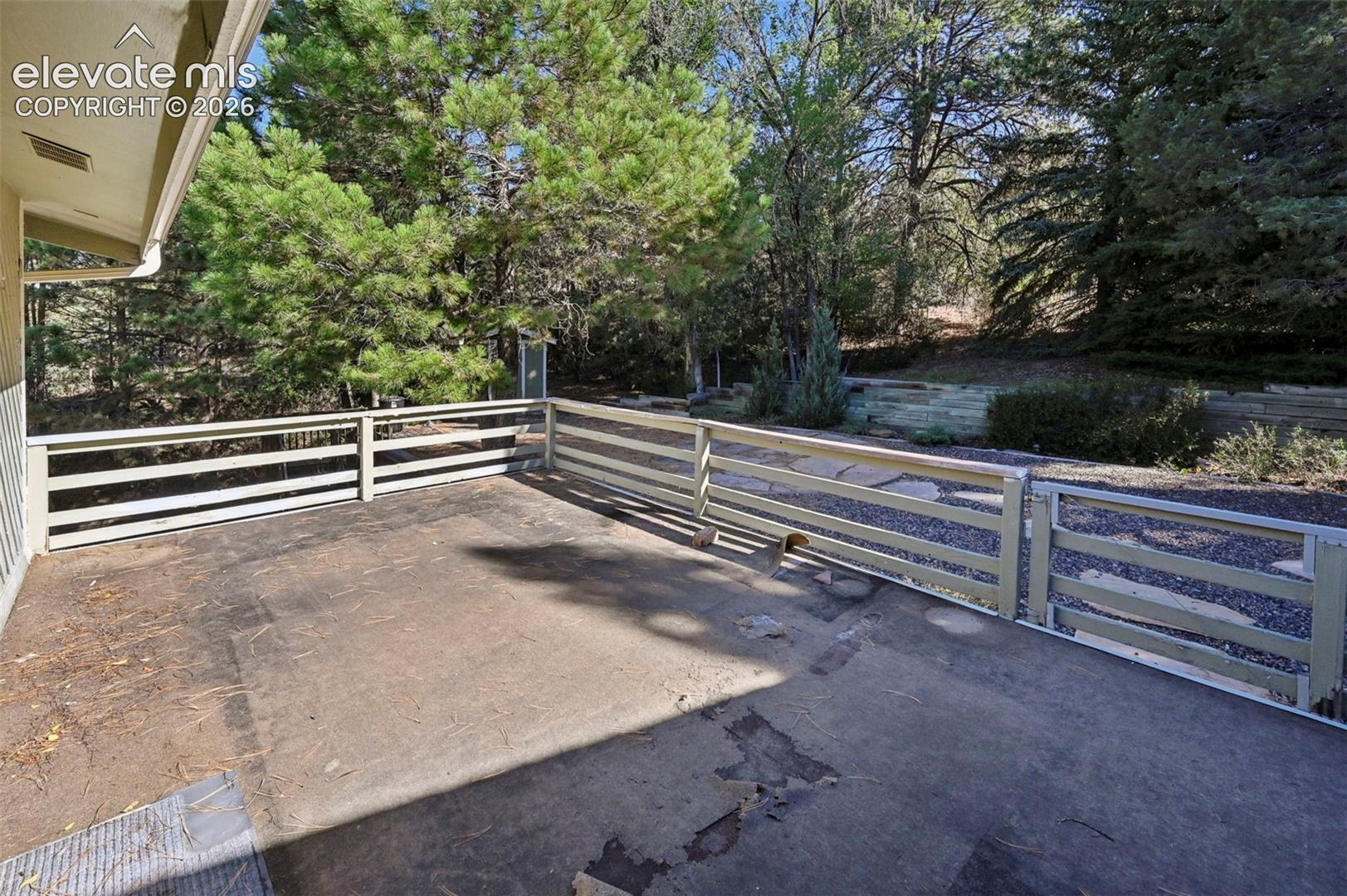 Concrete patio featuring a horizontal-slat wooden railing, tiered stone retaining walls, and mature evergreen trees