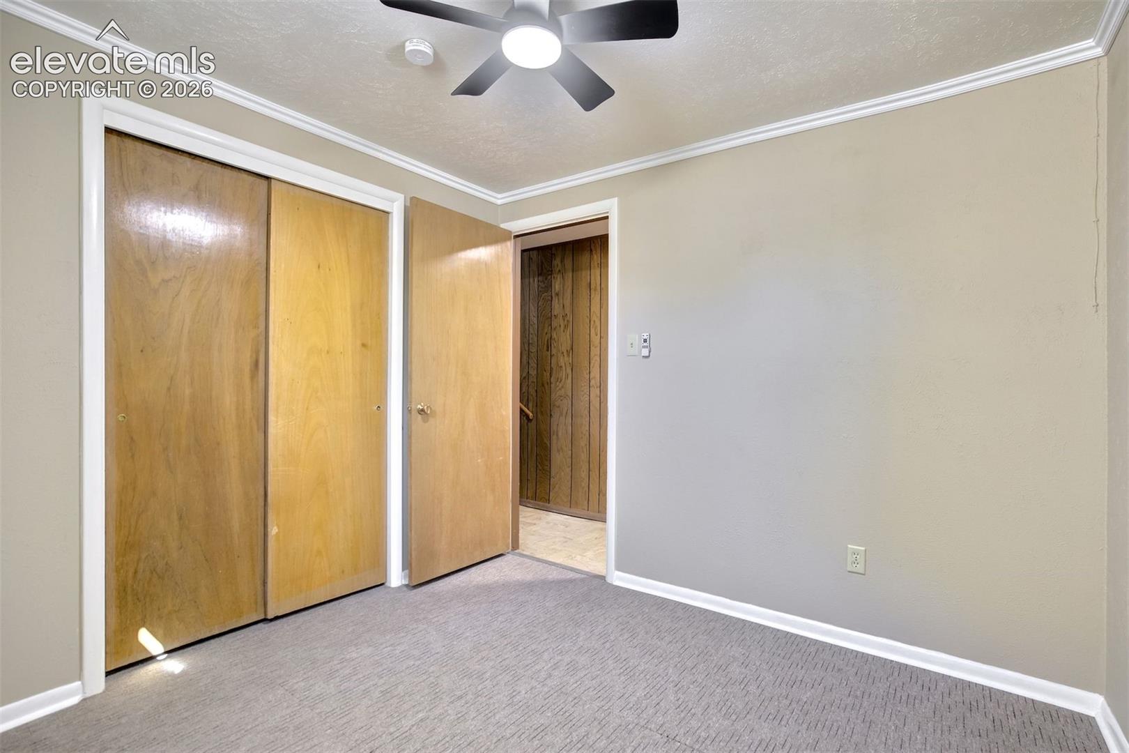 Room featuring wall-to-wall carpeting, wood-finish sliding closet doors, a neutral paint palette, crown molding, and a ceiling-mounted light fixture with fan
