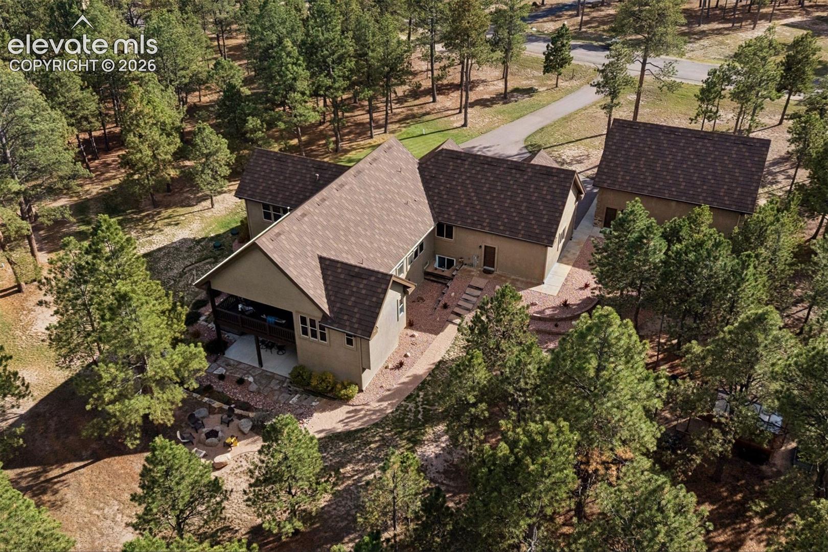 Aerial perspective featuring a multi-gabled exterior, an expansive paved patio, a stone fire pit, and a detached garage, all situated within a heavily wooded lot