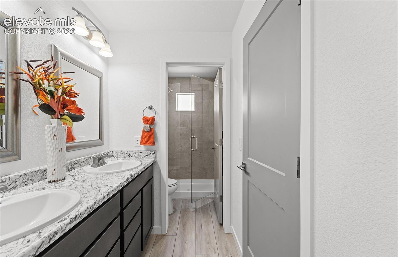 Dual-sink vanity with stone-pattern countertop, shaker-style cabinetry, wood-finish flooring, and glass-enclosed shower