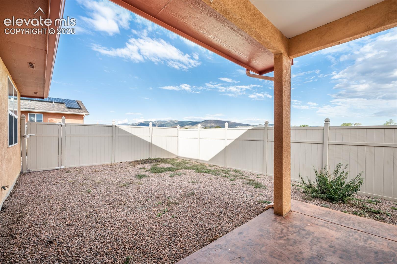Covered concrete patio featuring a perimeter of light-toned synthetic fencing, gravel grounds, and mountain range scenery
