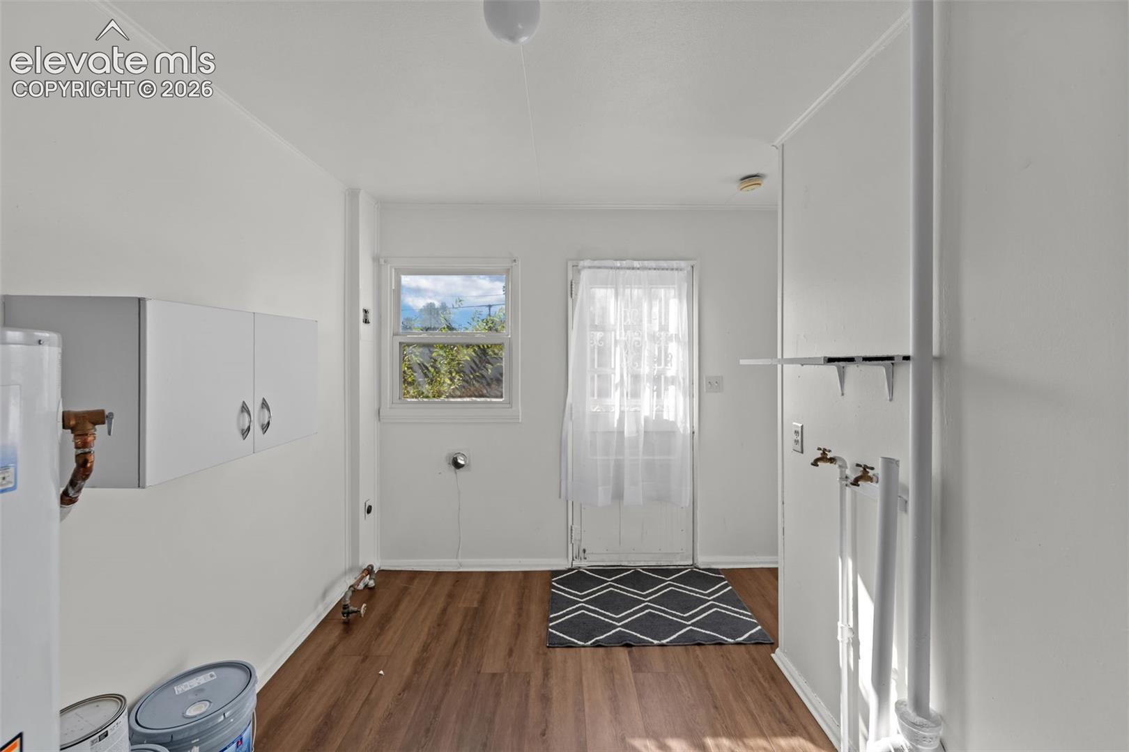 Utility area featuring wood-finish flooring, white wall-mounted cabinetry, a single-hung window, and dedicated plumbing connections