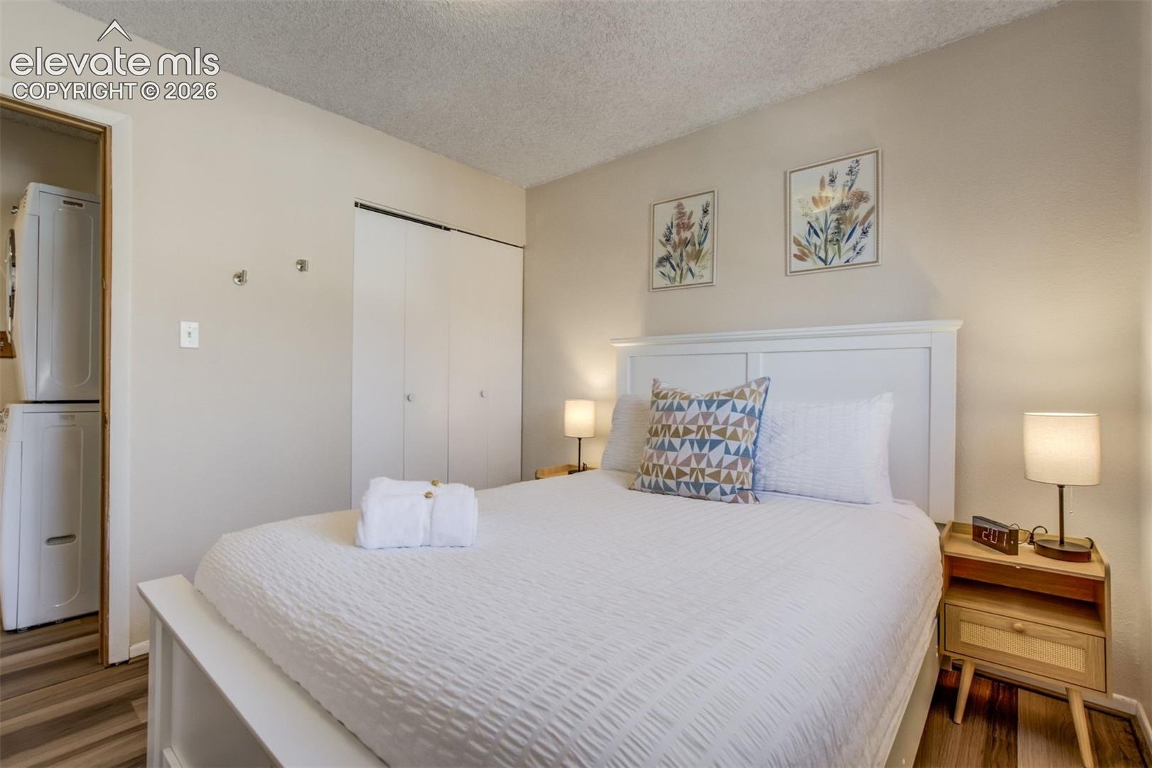 Stacked washer and dryer unit, wood-finish flooring, white paneled headboard, and sliding closet doors