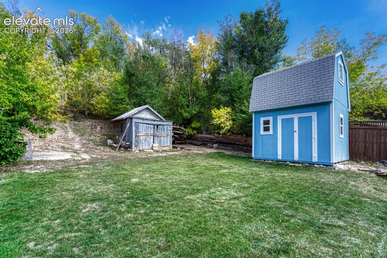 Expansive yard with green lawn, a bright blue storage shed with white trim, and a rustic storage structure