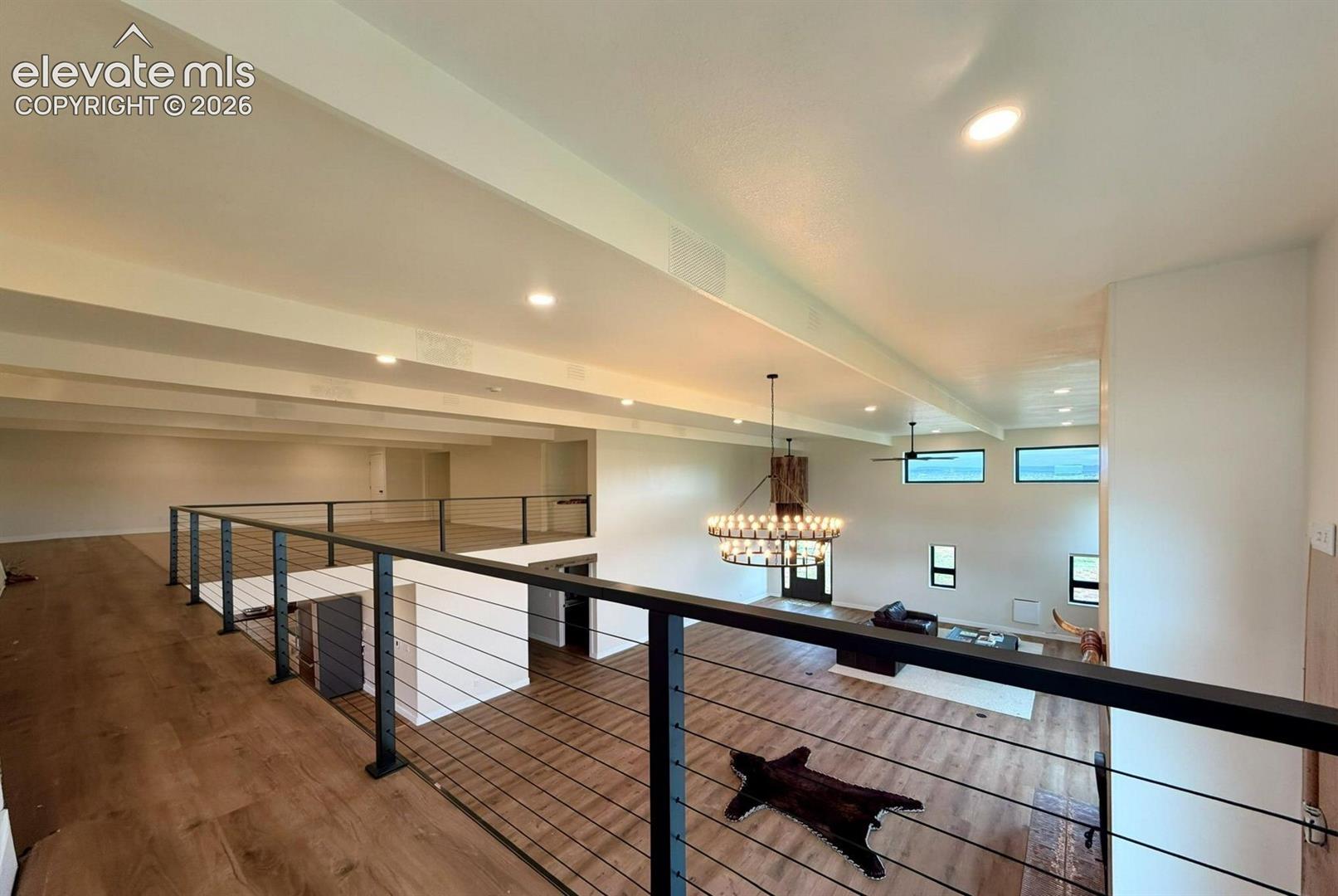 Open-concept mezzanine featuring wood-finish flooring, metal cable railing, exposed structural beams, and a large circular chandelier