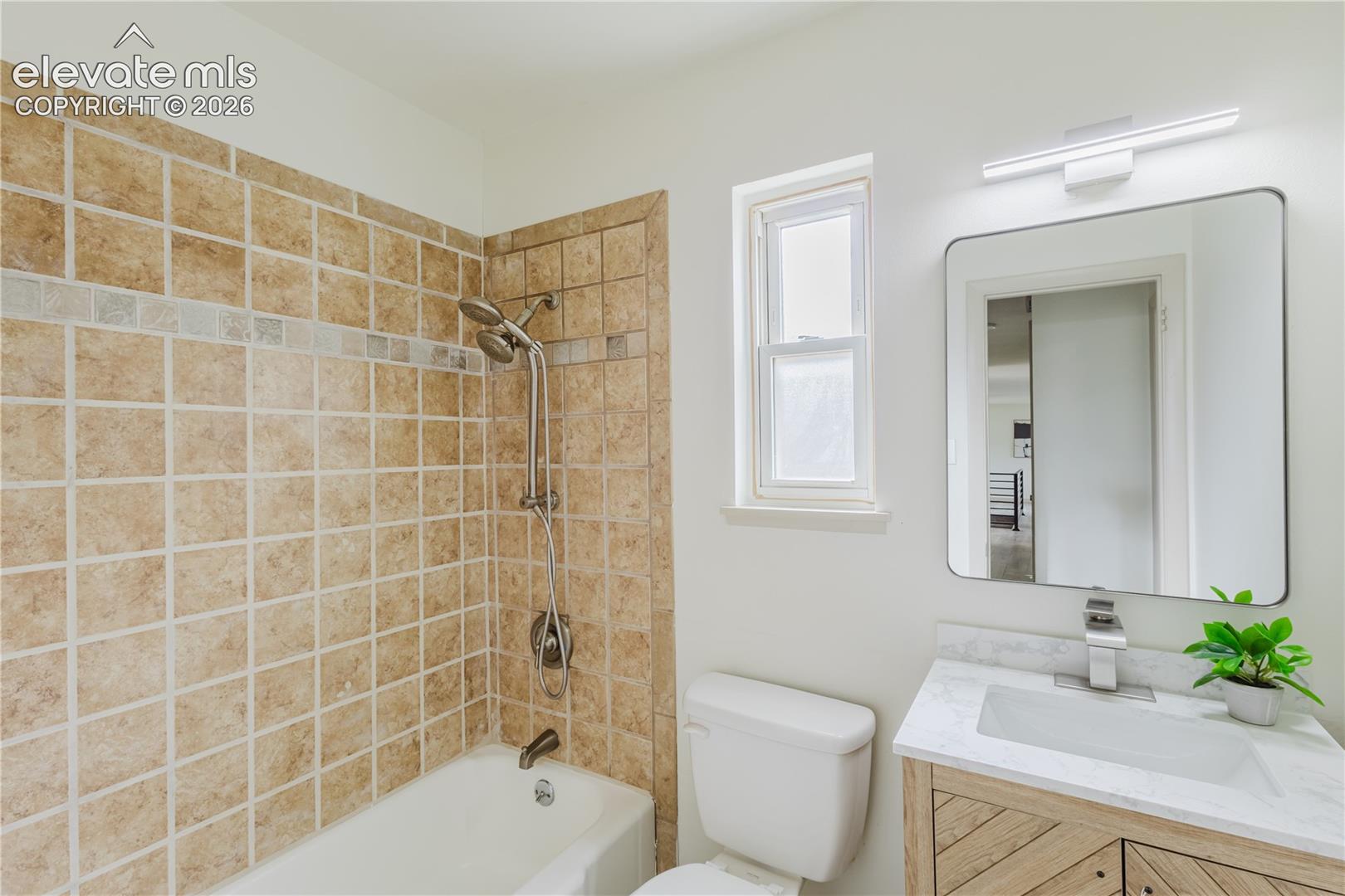 Bathroom featuring a tiled shower and bathtub combination with a multi-function showerhead, a single window, a toilet, a vanity with a rectangular basin, and a wall-mounted mirror with an overhead light fixture
