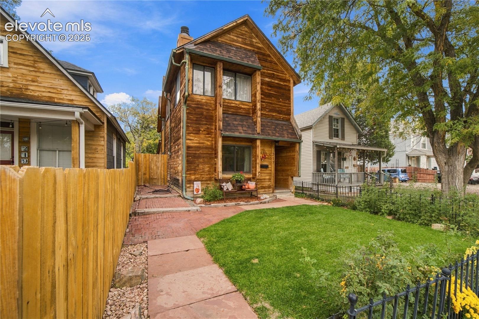 Multi-story exterior featuring natural wood siding, shingled roofing, a flagstone walkway, fenced front yard, and mature landscaping