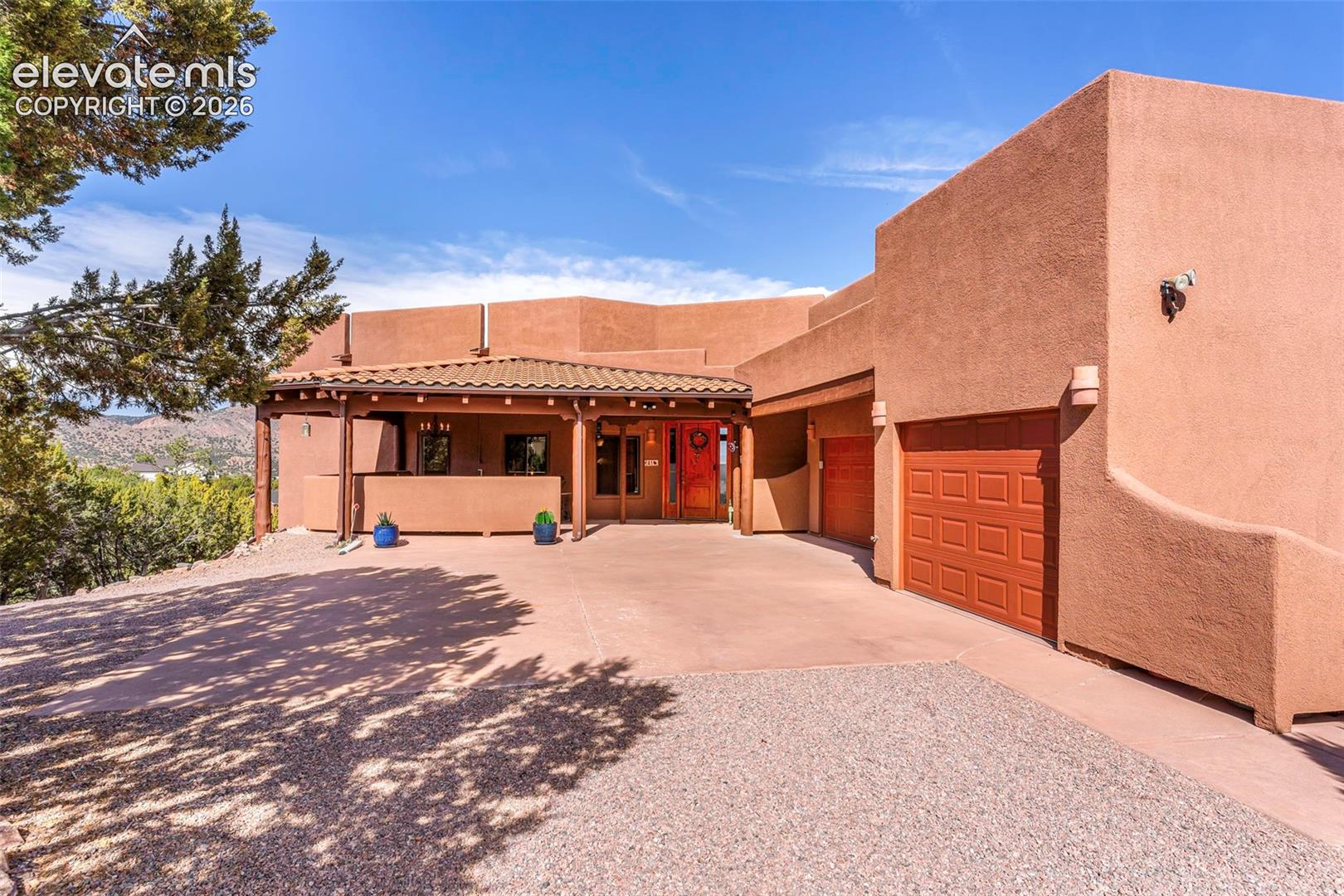Pueblo-style exterior featuring a stucco finish, covered entryway with wooden beam supports, terracotta tile roof, and a multi-car garage with paneled doors