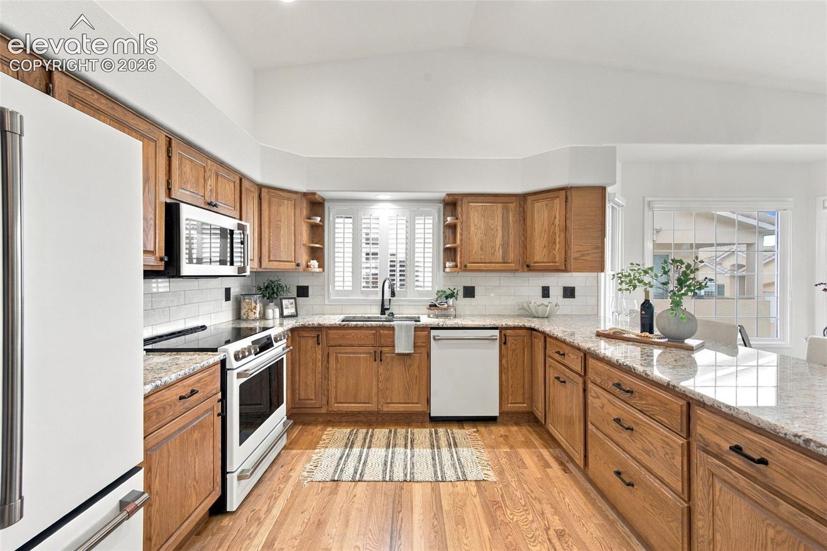 Kitchen featuring vaulted ceilings, light-toned wood-finish cabinetry, stone-surface countertops, subway tile backsplash, and wood-finish flooring