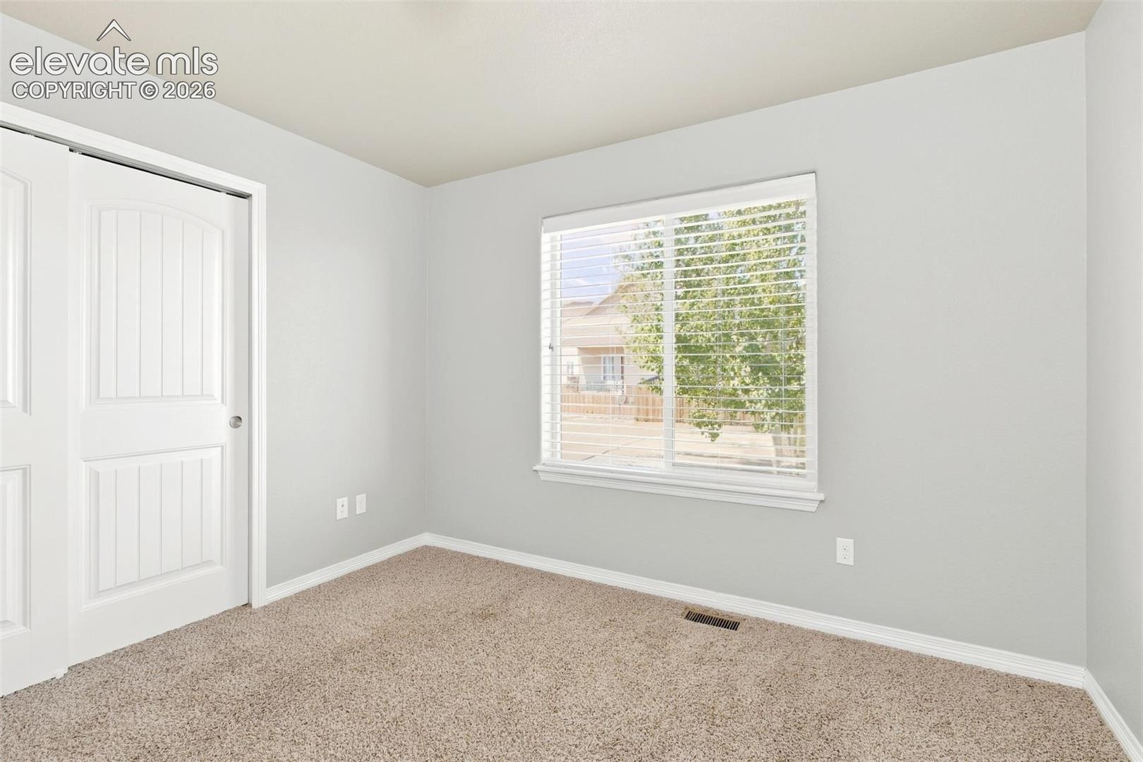 Neutral interior room featuring neutral-toned carpeting, light-colored walls, a sliding dual-panel closet door, and a single window fitted with horizontal blinds