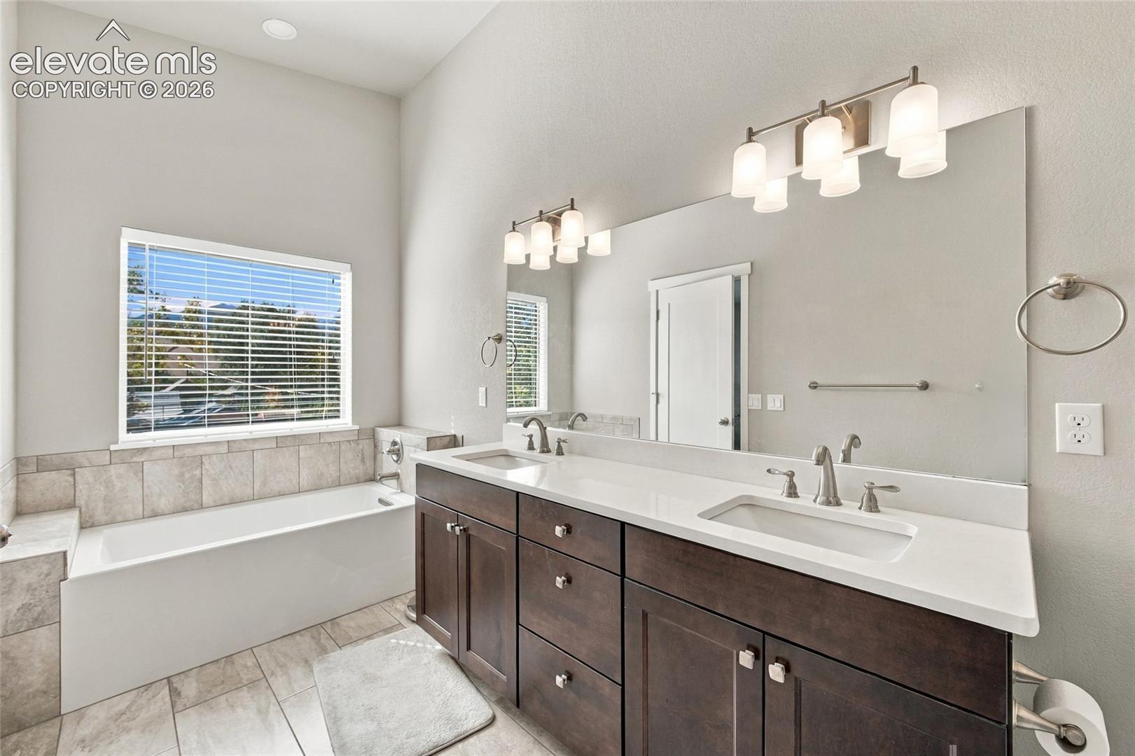 Dual-sink vanity with dark wood cabinetry and white countertop, large wall-mounted mirror, soaking tub with tile surround, and expansive window