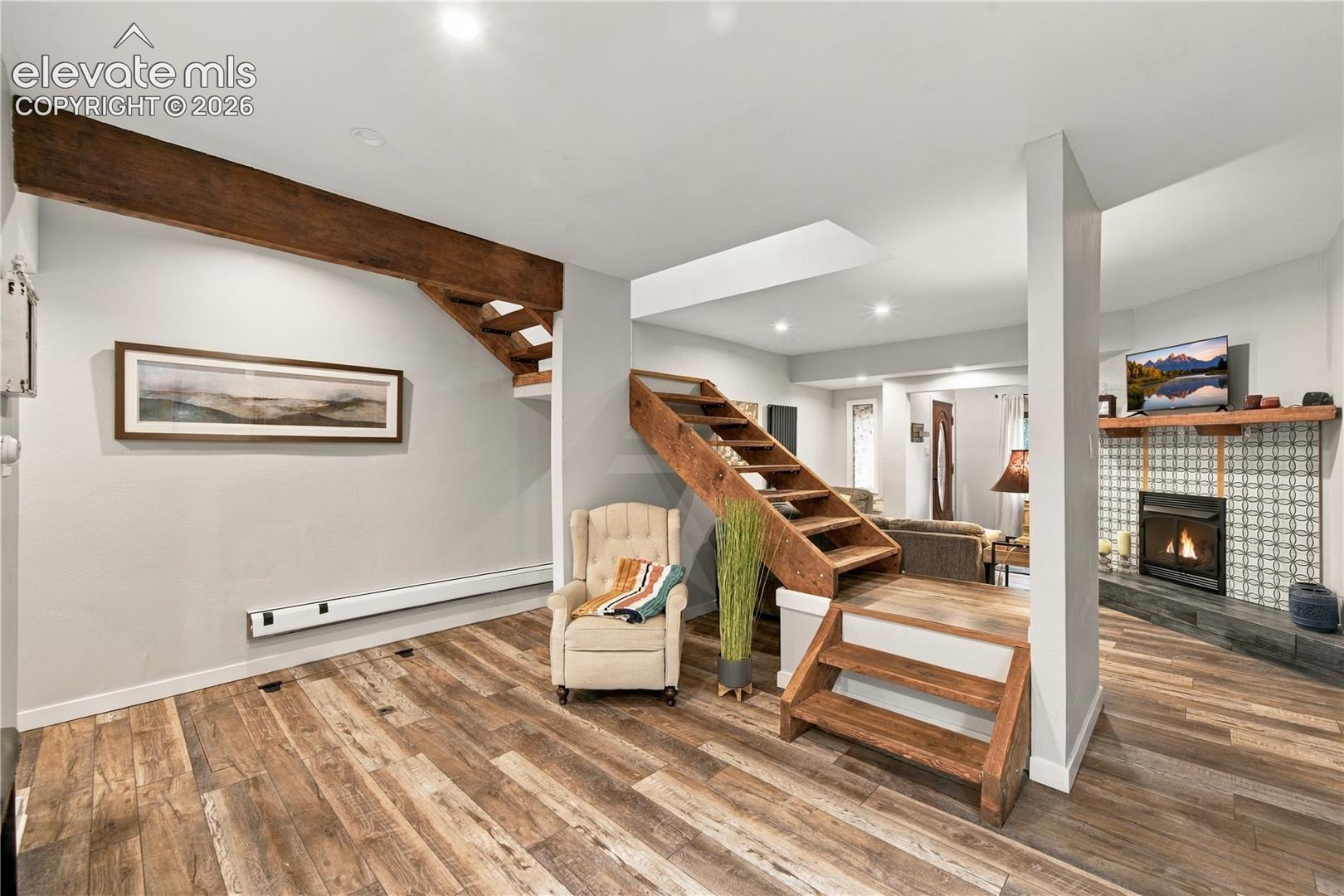 Open-concept interior featuring wood-finish flooring, exposed timber staircase, recessed ceiling lighting, and a fireplace with decorative tile surround and natural wood mantel