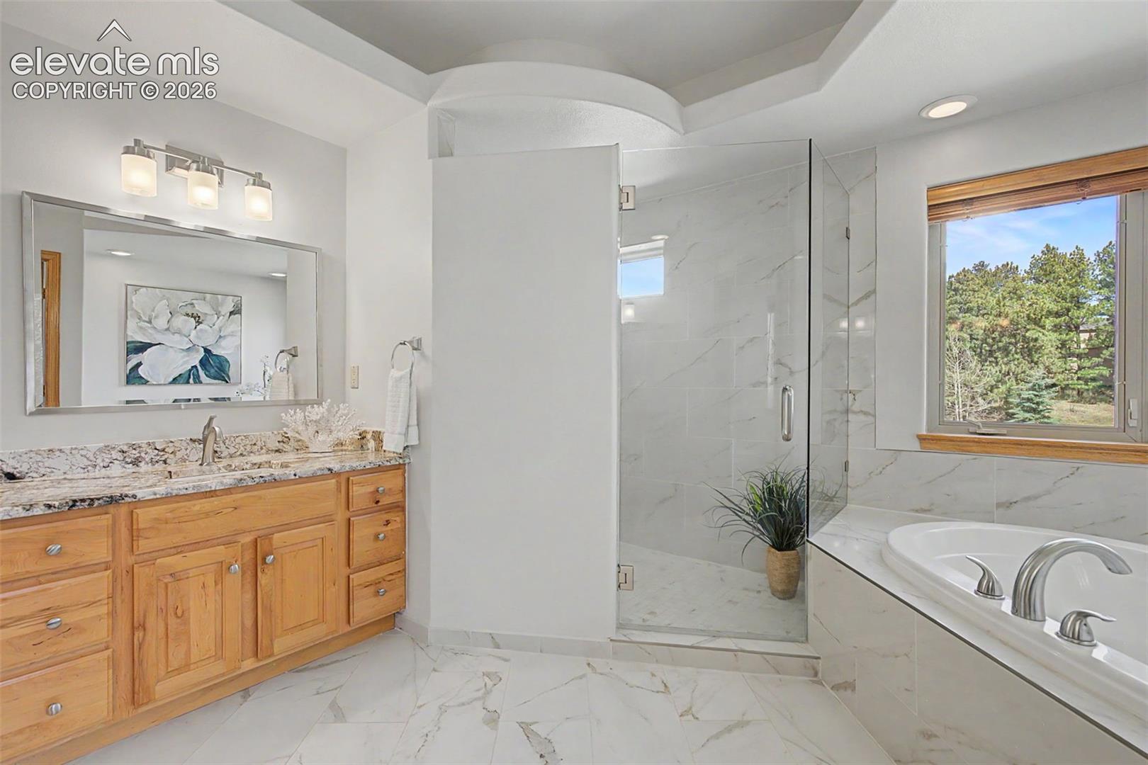 Bathroom featuring a spacious walk-in shower with clear glass enclosure and tiled walls, a separate soaking tub with tiled surround, a double vanity with stone countertop and wood cabinetry, and large format marble-look floor tiles