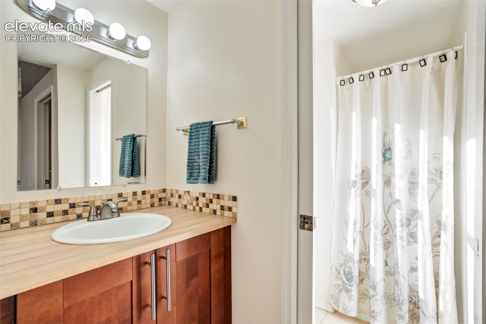 Bathroom featuring wood-finish cabinetry, a light-toned countertop, mosaic tile backsplash, oval undermount sink, and wall-mounted vanity lighting