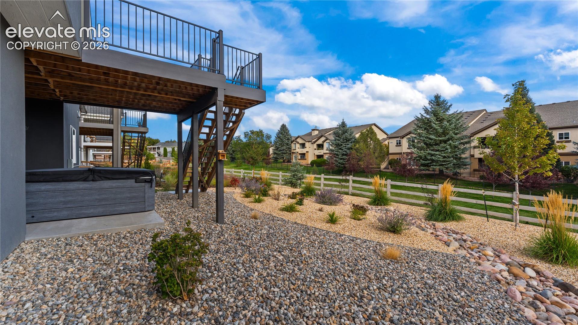 Covered concrete patio featuring a built-in spa, elevated wood-frame deck with metal railing, xeriscaped rock garden, and perimeter white rail fencing