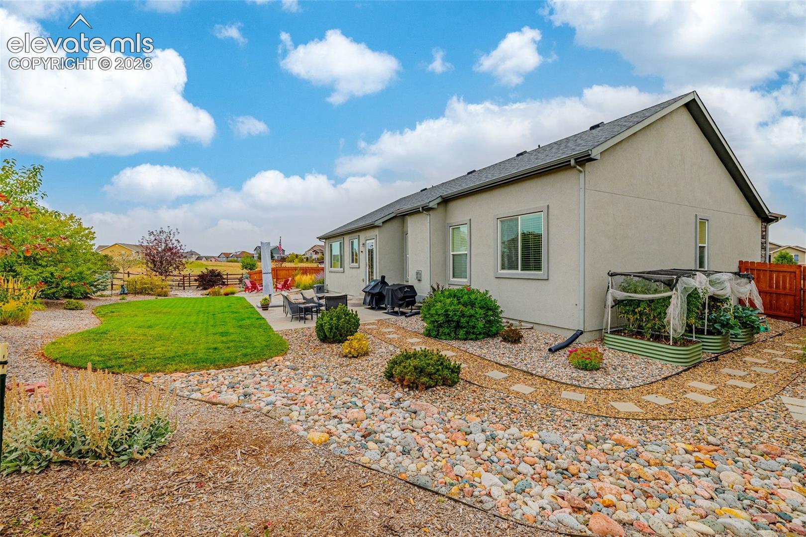 Rear exterior featuring a concrete patio, xeriscaped grounds with river rock, a manicured lawn, and raised garden beds