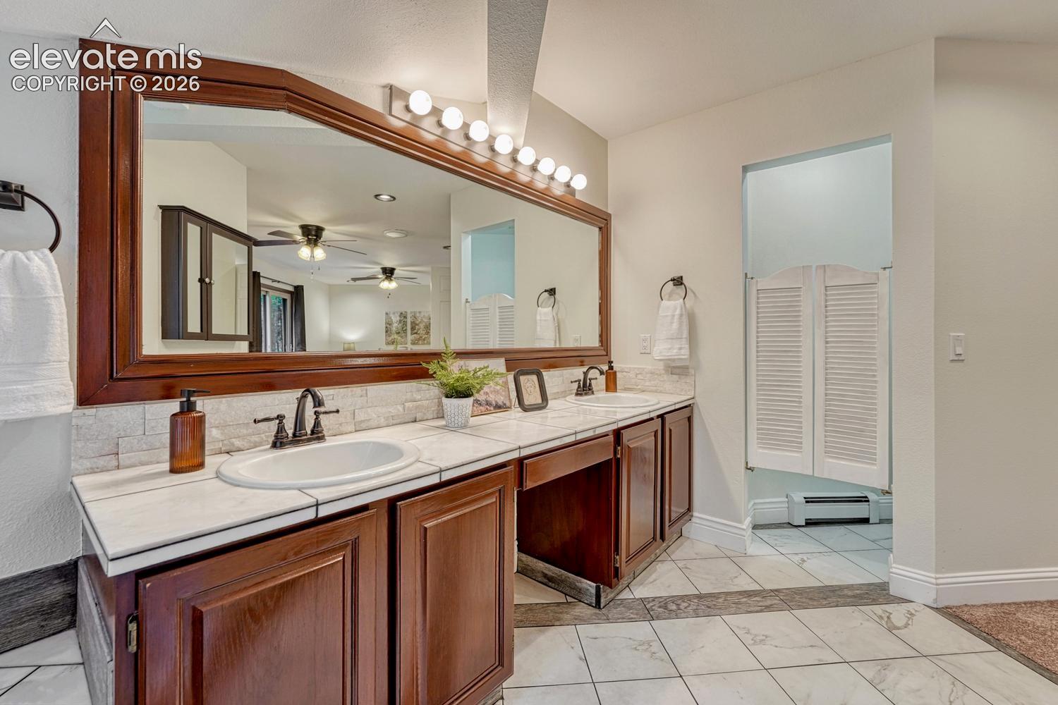 Double-sink vanity featuring wood cabinetry, tile countertops, natural stone-style backsplash, integrated lighting, and a large framed mirror