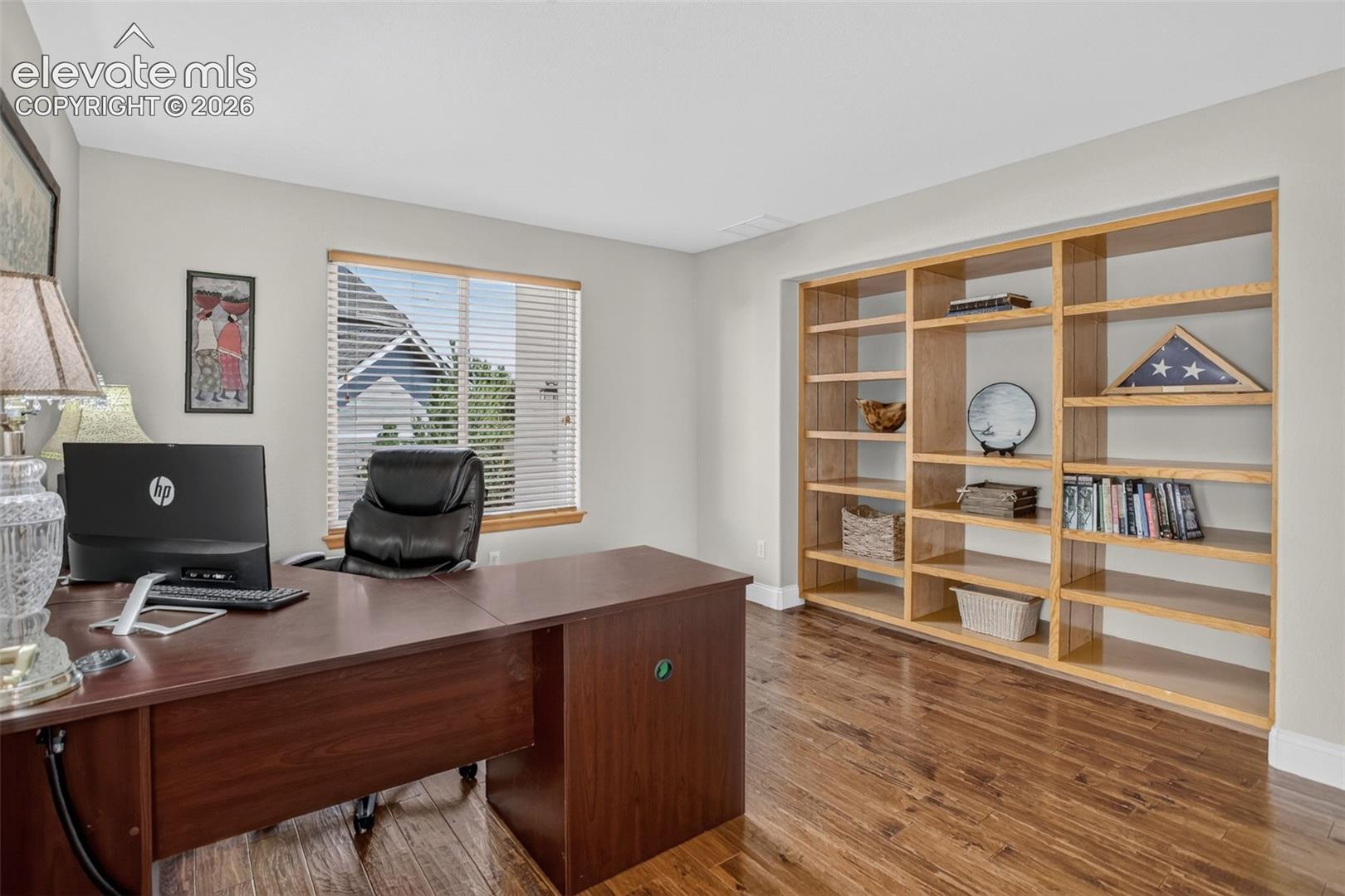 Hardwood-finish flooring complementing built-in shelving, a large window with blinds, and white baseboards