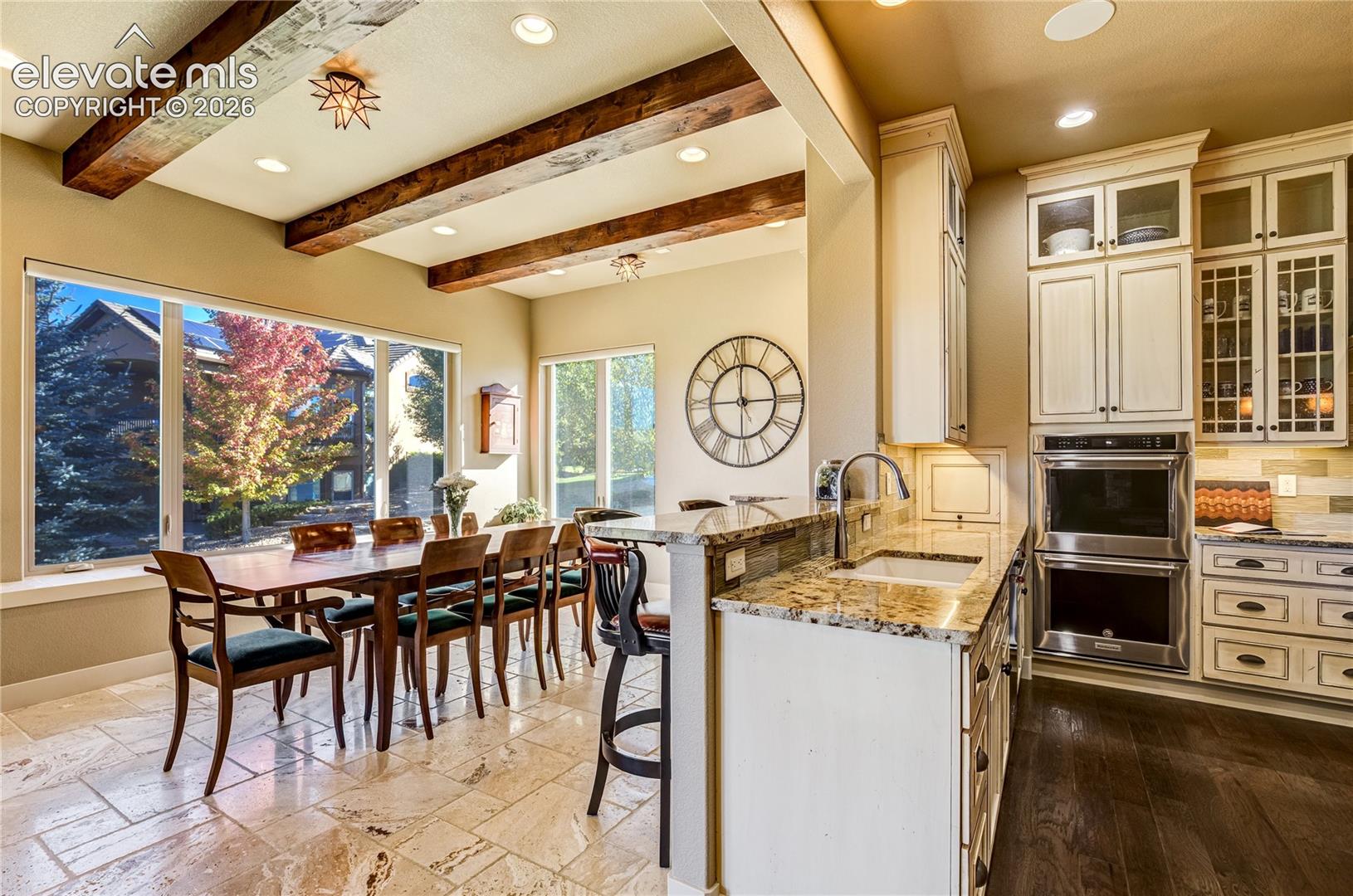 The beautifully appointed kitchen flows into the light-filled garden room, where stained ceiling beams, travertine tile + a custom dry bar create an inviting setting for casual dining + effortless entertaining.