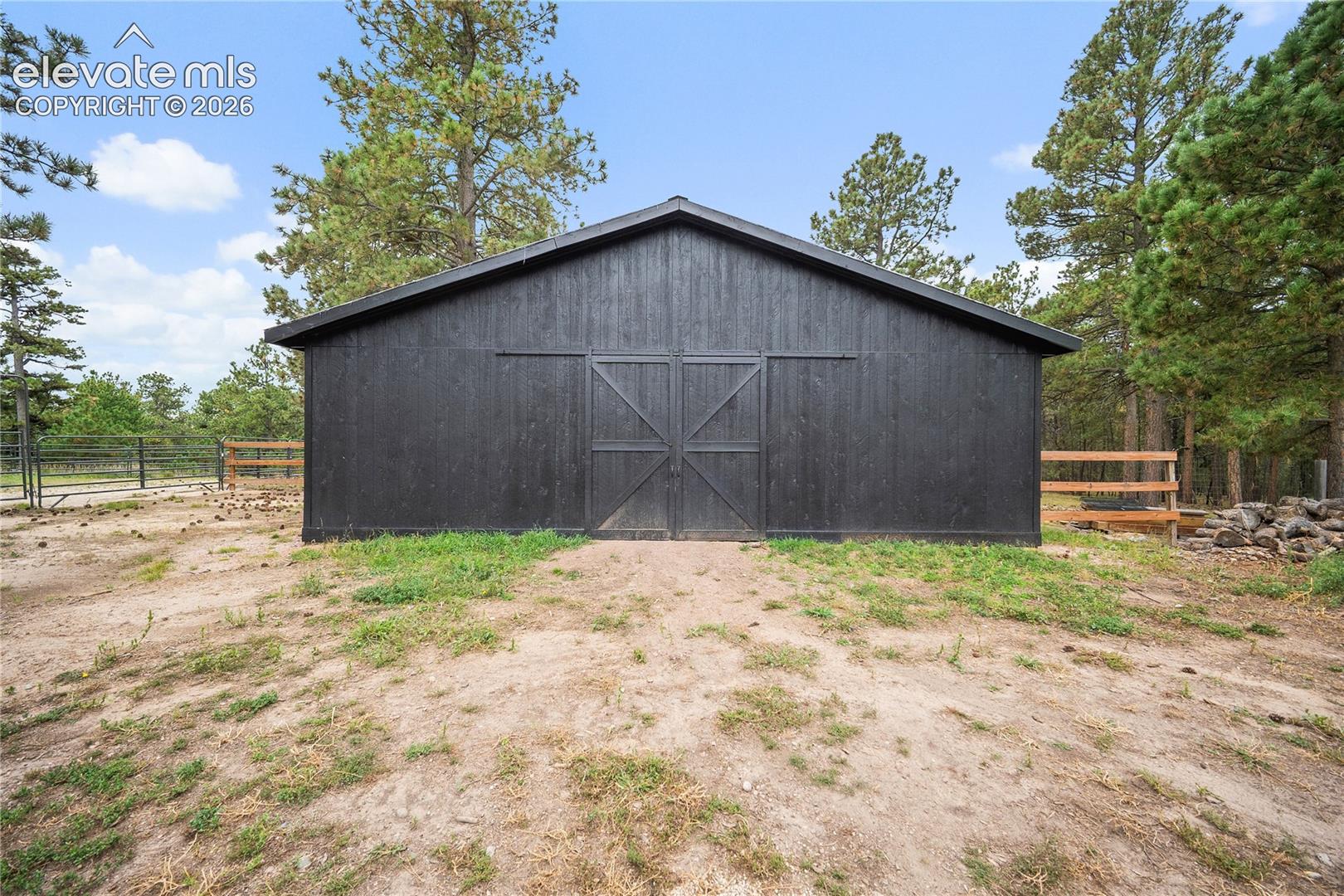 Black-painted wood-sided exterior building featuring double sliding barn-style doors and a gabled roof, situated on a lot with mature pine trees and wood-rail fencing