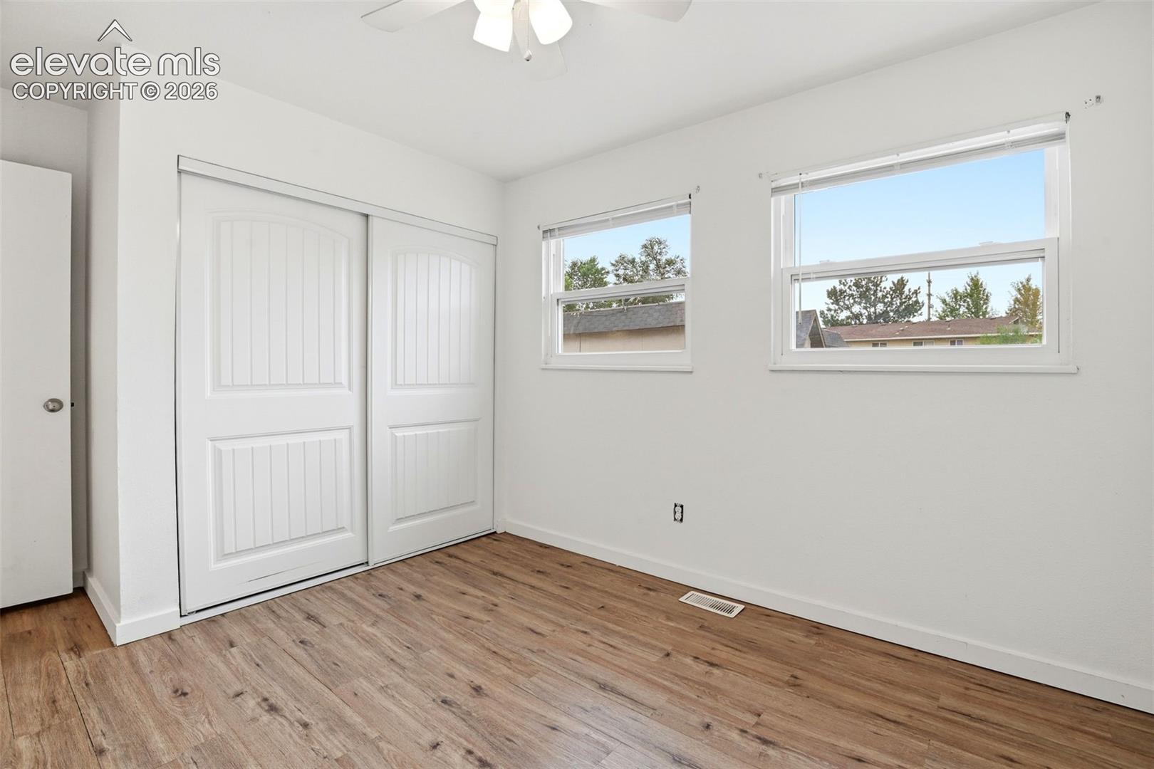 Wood-finish flooring, two large windows, white walls, built-in bi-fold closet doors, and a ceiling fan