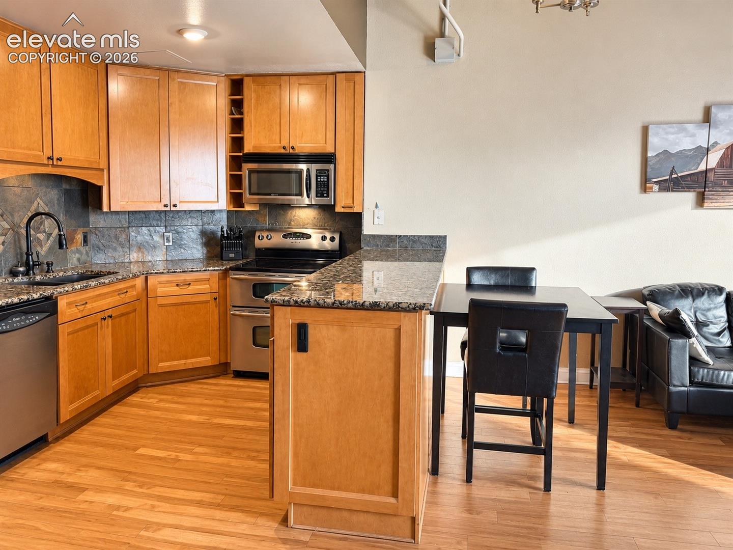 Kitchen featuring light wood-finish cabinetry, stainless steel appliances, speckled stone countertops, and a gray tile backsplash