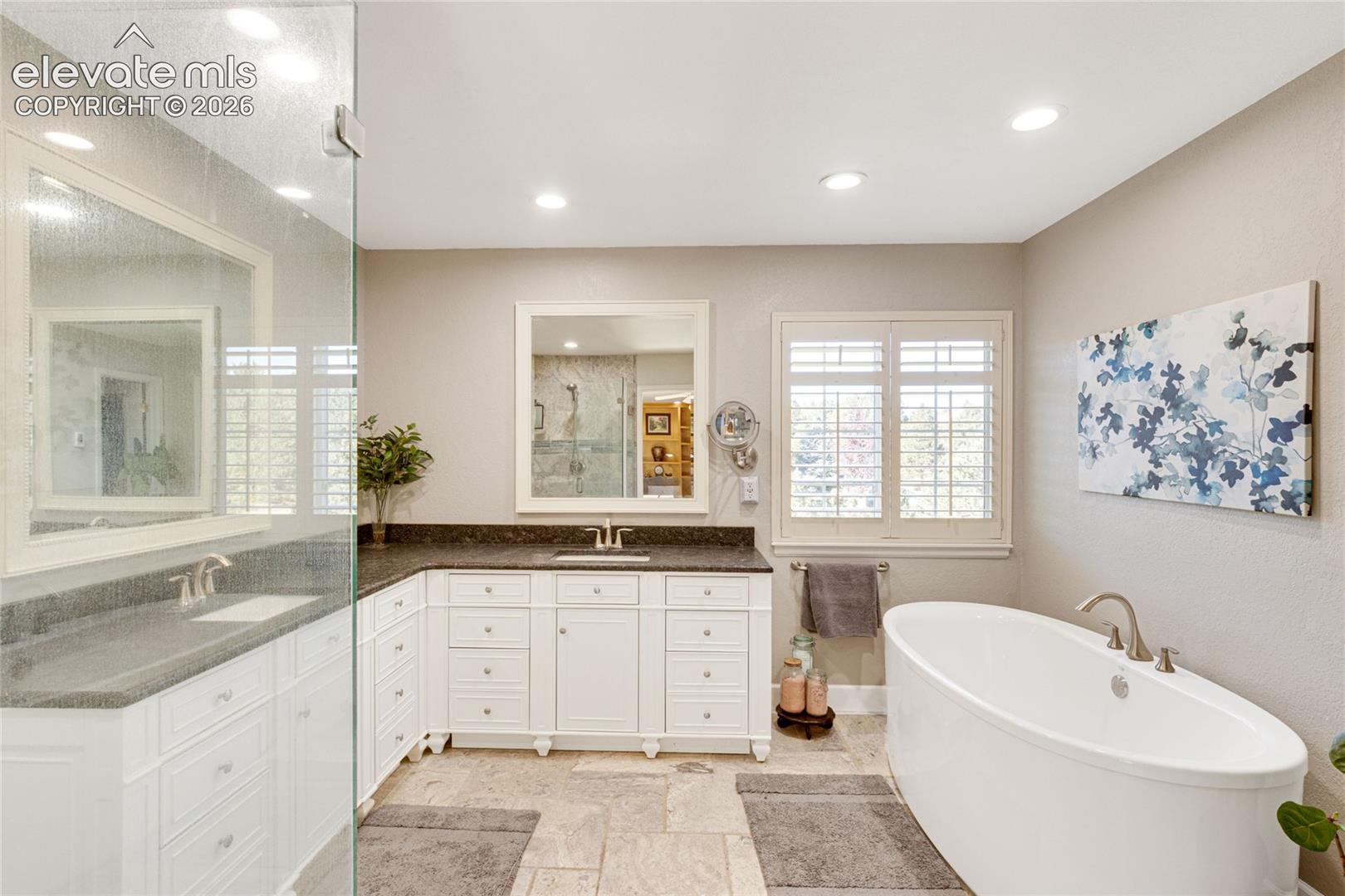 L-shaped white cabinetry with dark stone countertops, dual vanity sinks, freestanding soaking tub, recessed lighting, and tile flooring