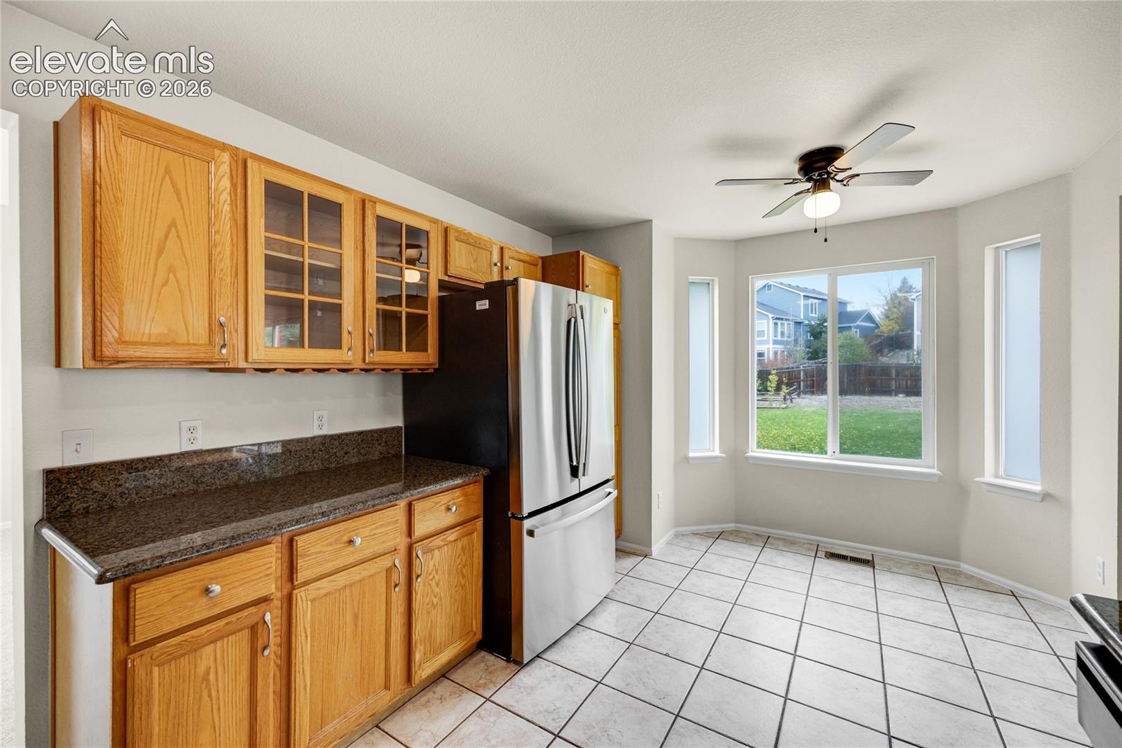 Kitchen featuring natural wood cabinetry with glass-front uppers, stone-finish countertops, stainless steel refrigerator, tile flooring, and a multi-window bay window configuration