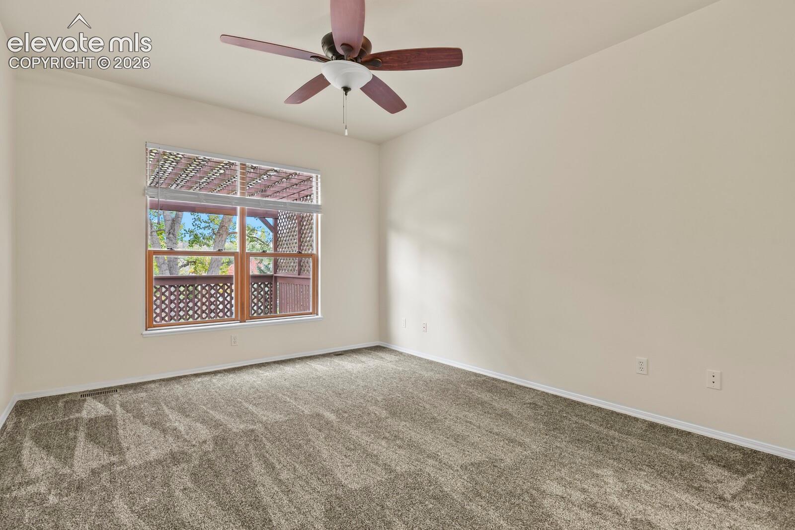 Neutral-toned interior room featuring wall-to-wall carpeting, a multi-blade ceiling fan with integrated lighting, and a large window with wooden trim