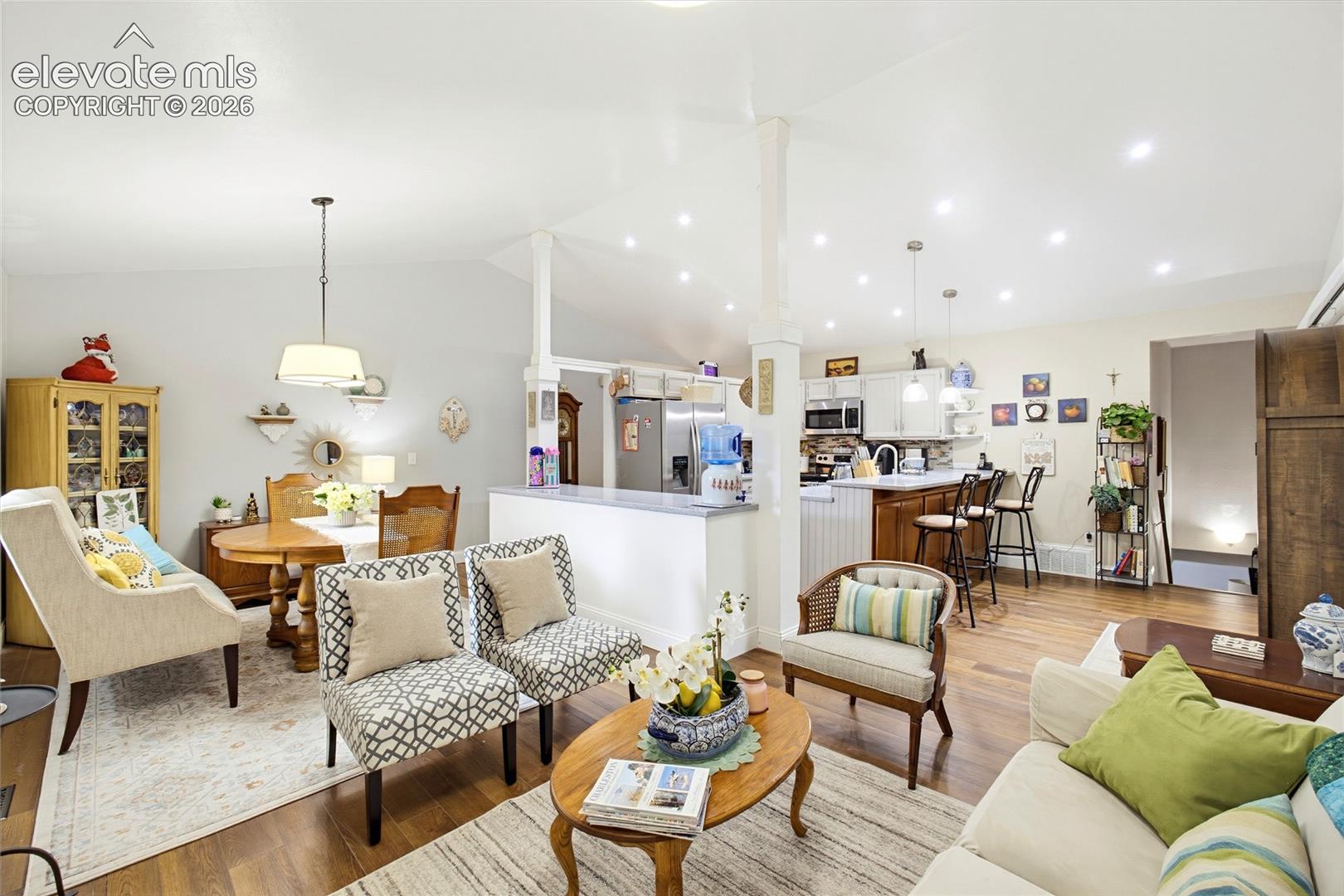 Open-concept living area featuring wood-finish flooring, vaulted ceilings with recessed lighting, and a kitchen with white cabinetry and a breakfast bar