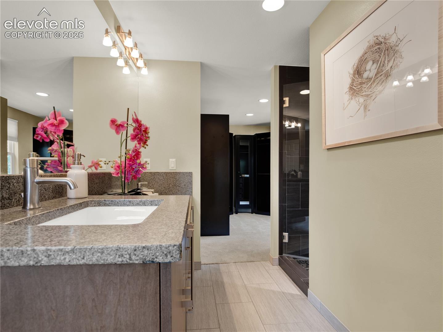 Vanity featuring a stone countertop with an integrated basin, multi-bulb light fixture, expansive wall-mounted mirror, and glass-enclosed shower with dark tile surround