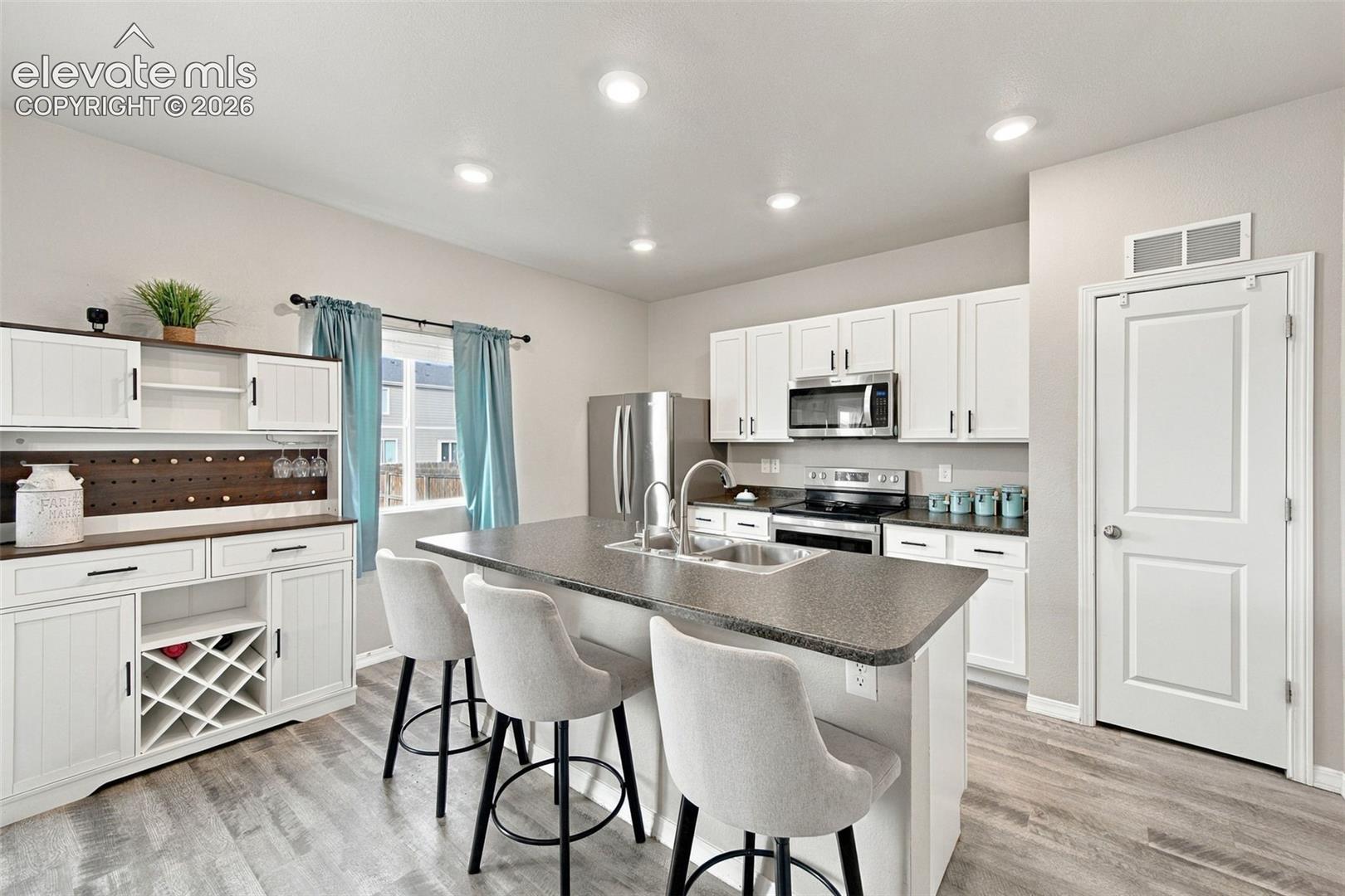 Kitchen featuring white cabinetry, a center island with a dual-basin stainless steel sink, wood-finish flooring, recessed lighting, and built-in wine storage