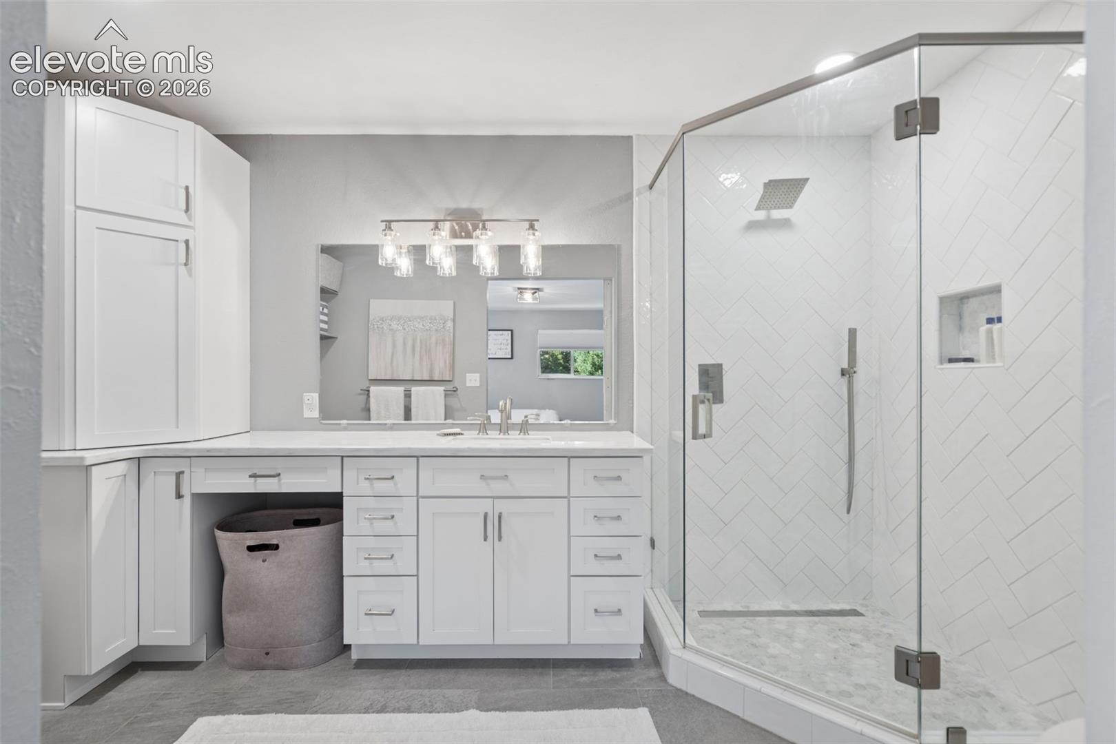 White shaker-style cabinetry with extensive counter space, large-format mirror, five-light fixture, glass-enclosed shower with herringbone tile walls, and rainfall showerhead