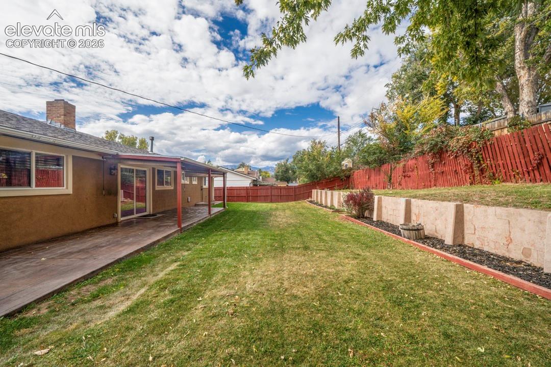 Backyard featuring a covered concrete patio, tiered retaining walls, manicured lawn, and wooden perimeter fencing