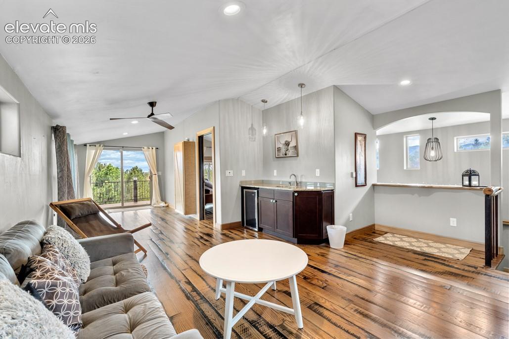 Vaulted ceiling with recessed lighting, wood-finish flooring, built-in wet bar with dark wood cabinetry, ceiling fan, and sliding glass door to a deck