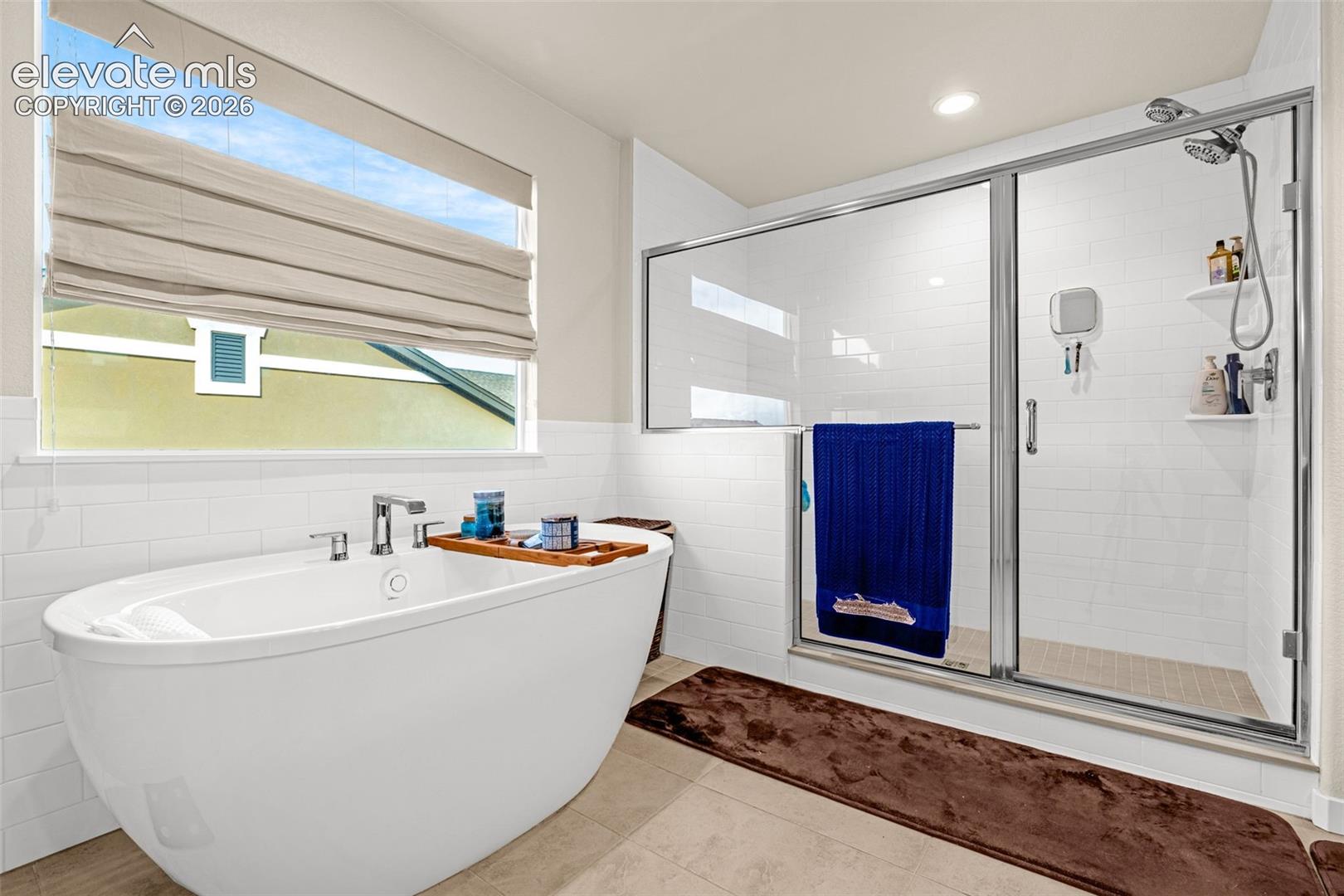Freestanding soaking tub with chrome fixtures, glass-enclosed shower with white subway tile surround, and large window with integrated blind