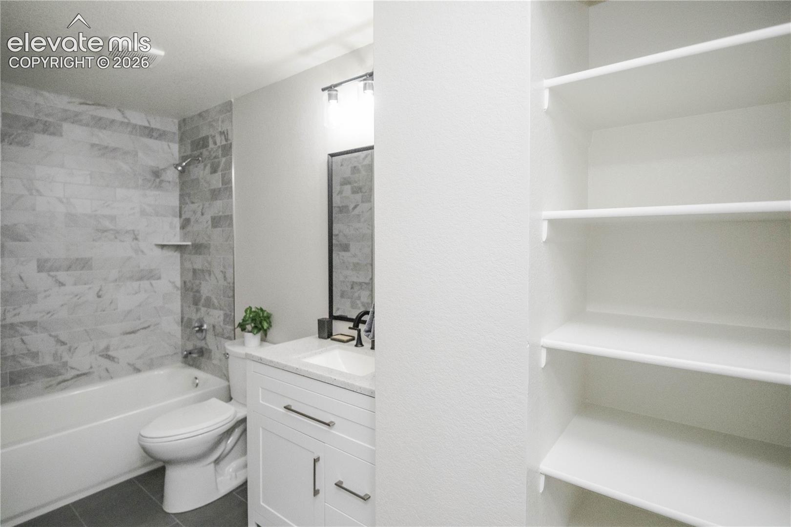 Bathroom featuring a shower-tub combination with stone-tile surround, white vanity cabinet with a light-toned countertop, wall-mounted light fixture, and built-in shelving